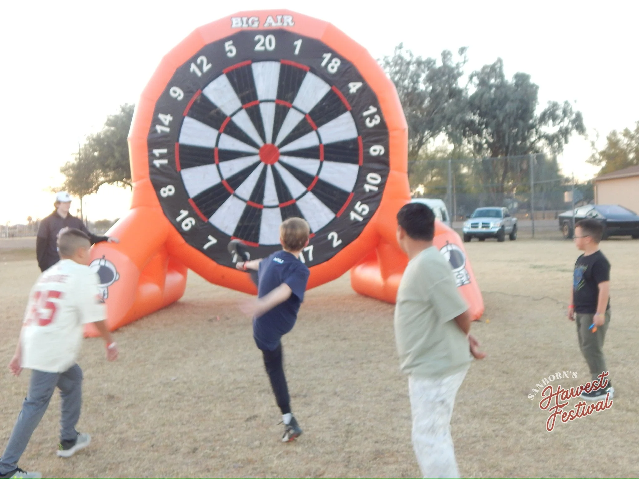 Group of children playing near an inflatable outdoor dartboard at a festival, with trees and parked cars in the background.