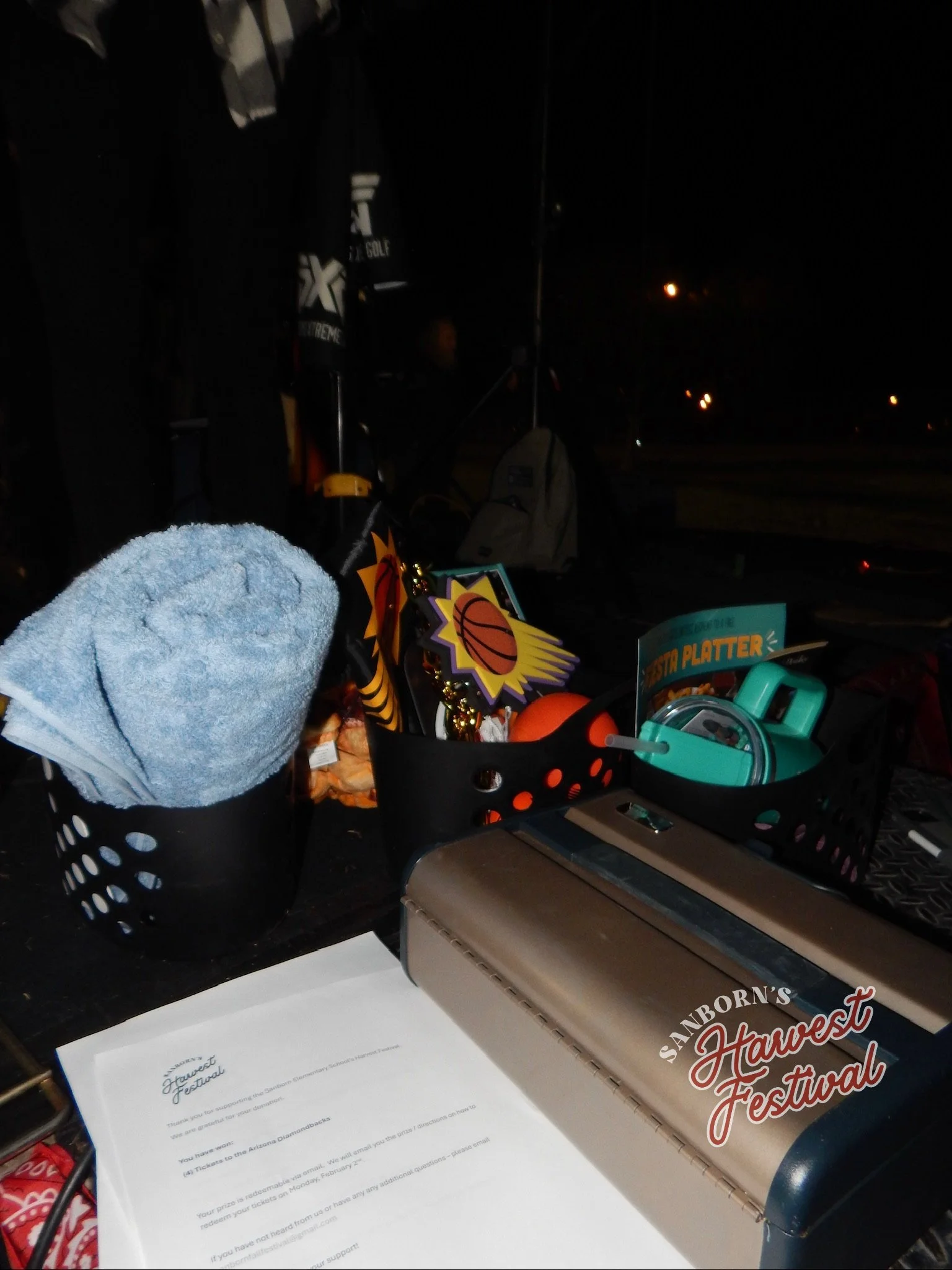 A desk in a dark room with a black shopping basket filled with various items, a paper titled "Sandborn's Harvest Festival," and a black device, possibly a portable fan. The background shows a person with a backpack, some illuminated objects, and a da