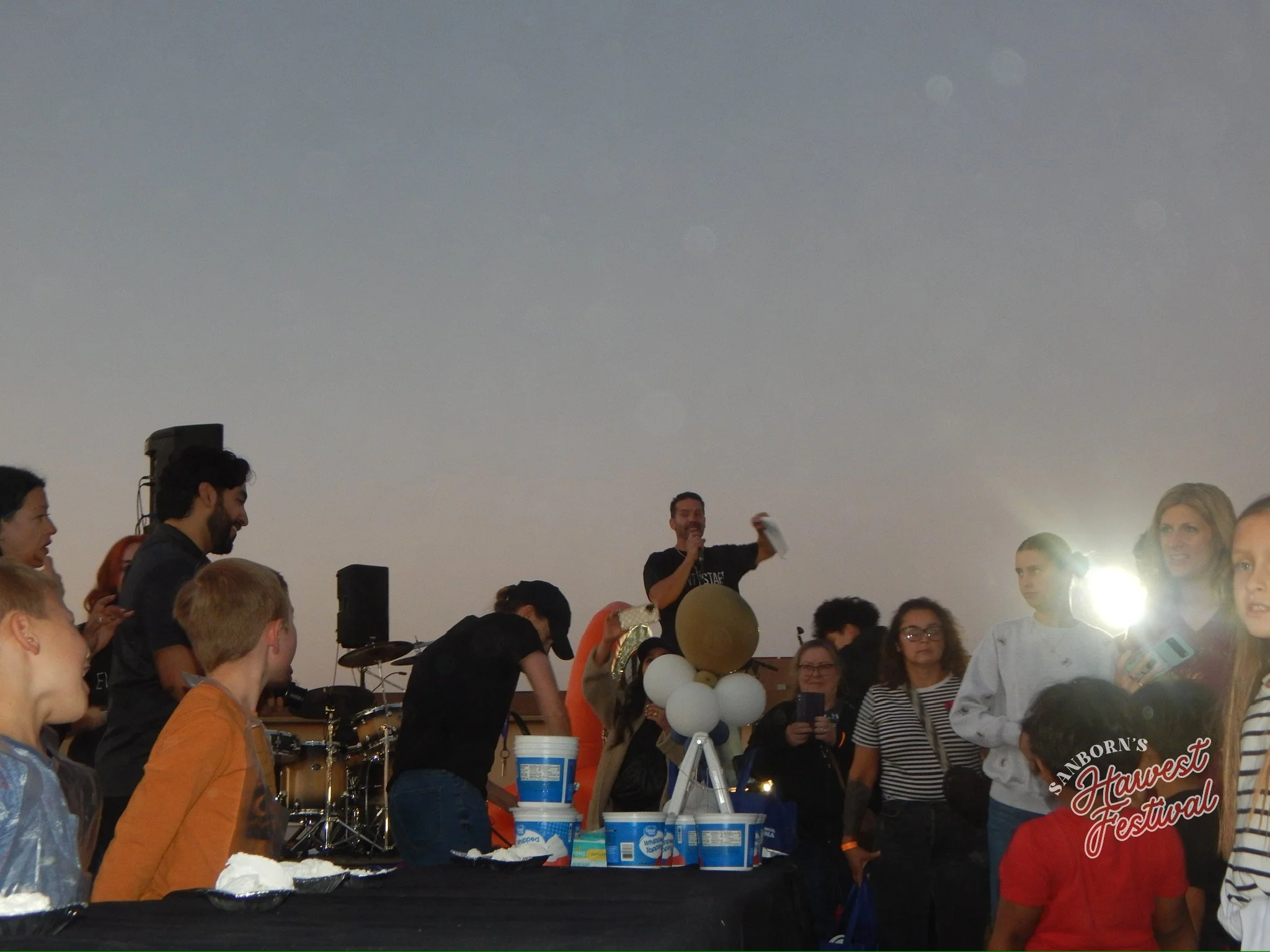A group of people gathered indoors at a festival, some wearing rain ponchos, with balloons, a cake, and a banner that reads 'Sanborn's Harvest Festival' in the background.