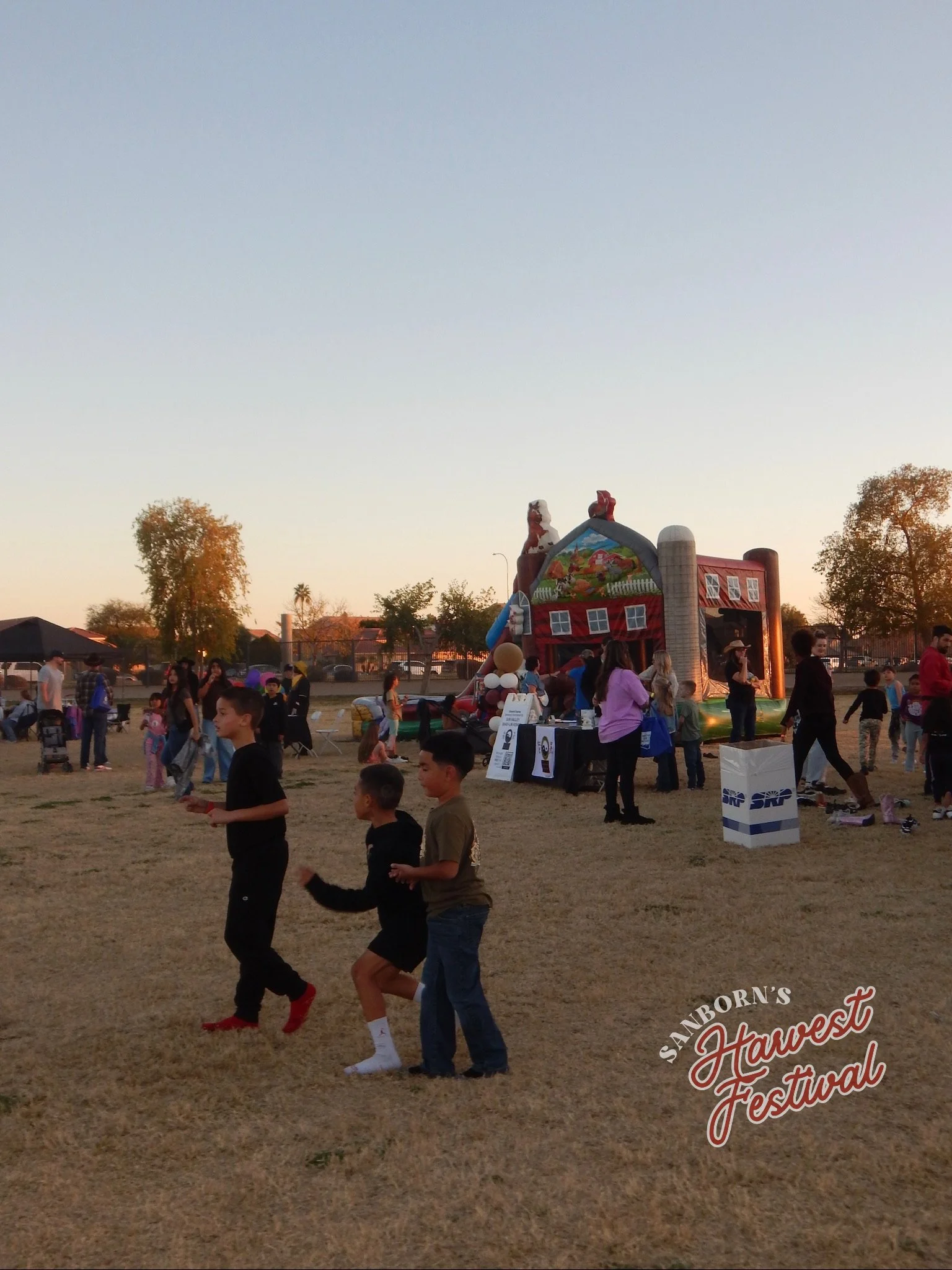 People walking and enjoying the outdoors at the Sanford's Harvest Festival, with a festive inflatable structure and trees in the background during sunset.