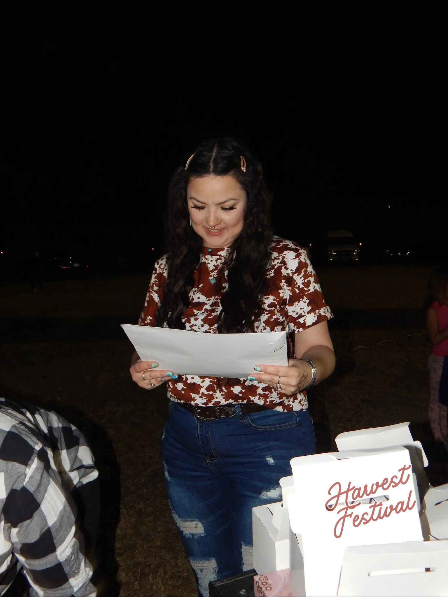 A young woman with long dark hair, wearing a cow print shirt, looking at papers at night outdoors during a Harvest Festival event.