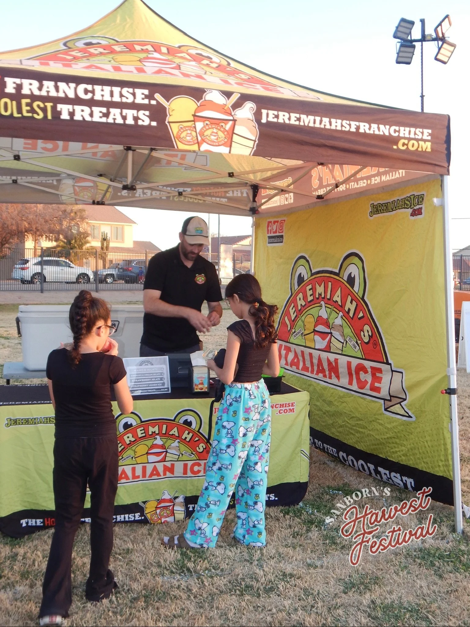 Children at an ice cream stand with a yellow and brown tent with colorful ice cream cone graphics and text advertising Italian ice. The background shows a parking lot with cars and houses, and a few adults are assisting the children.