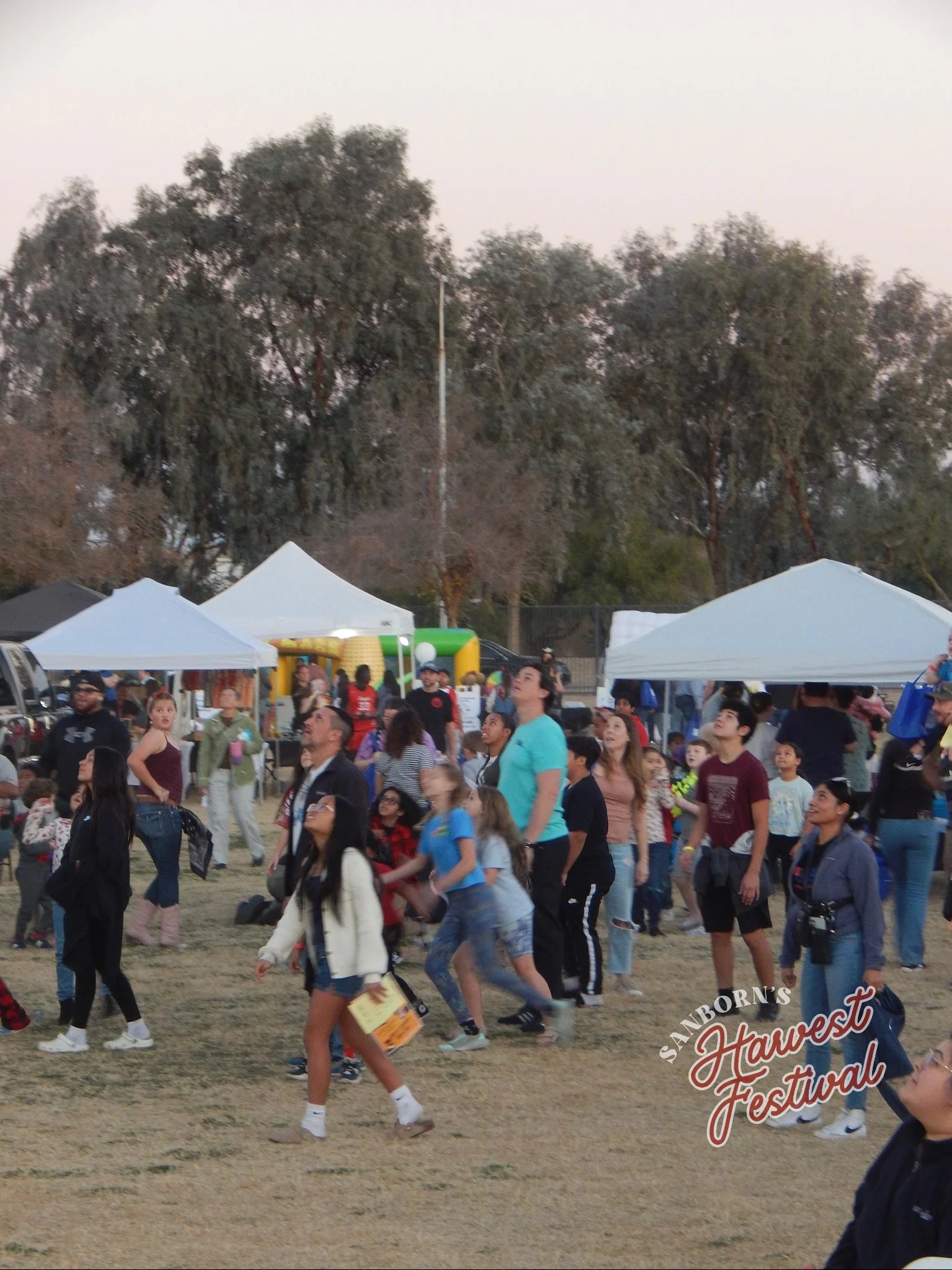 Crowd of people attending the Sanborn's Harvest Festival outdoors during daytime, with white tents and trees in the background.