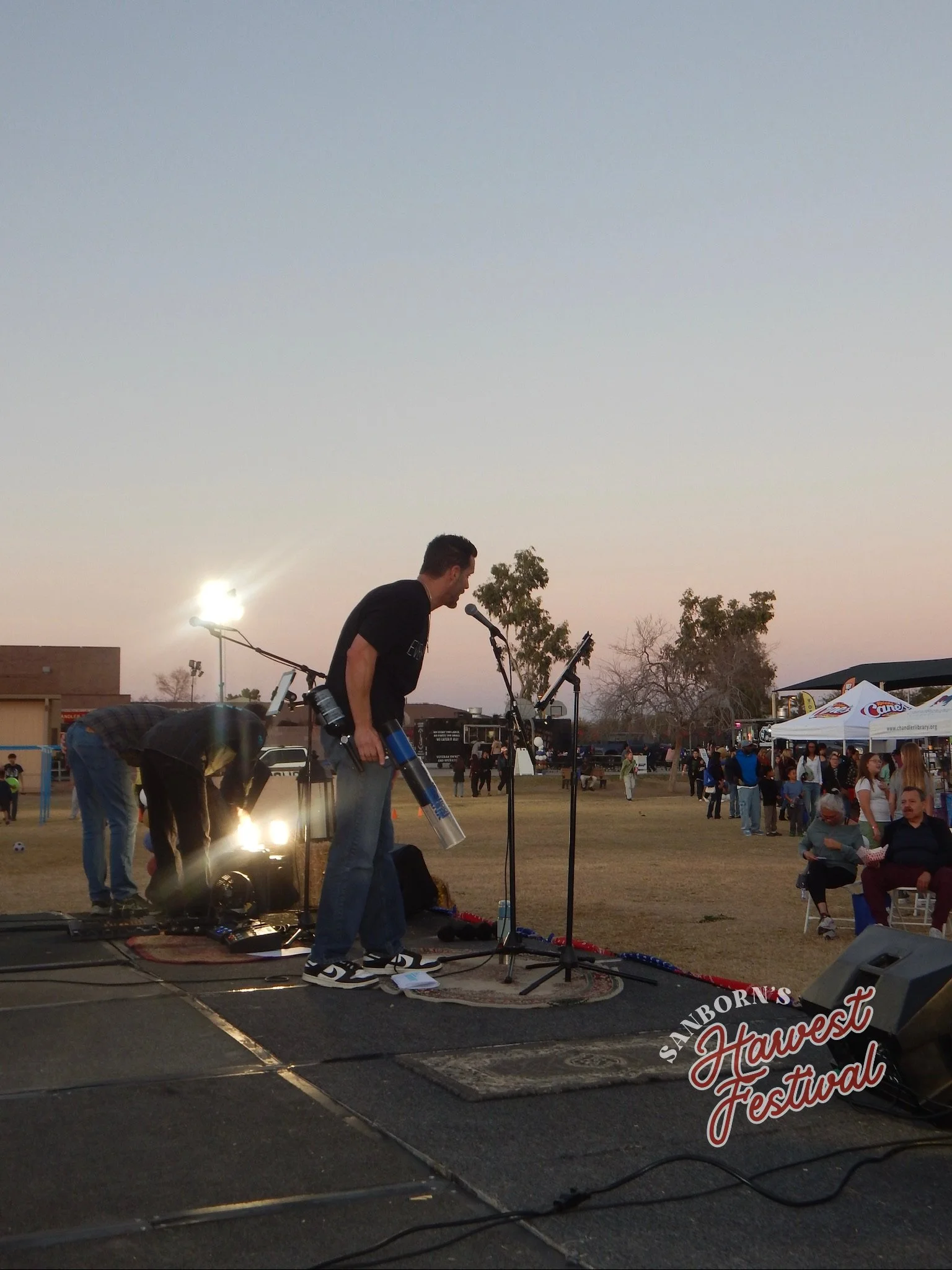 A man setting up equipment on a stage at sunset during a harvest festival, with people gathering in the background and a tent with a sign that reads 'Santa Barbara's Harvest Festival.'