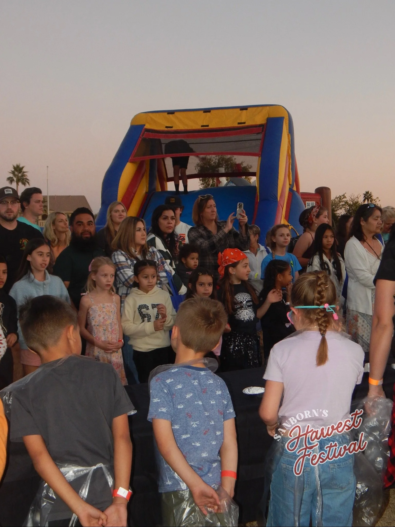 Group of children and adults at an outdoor harvest festival, with a bounce house in the background.
