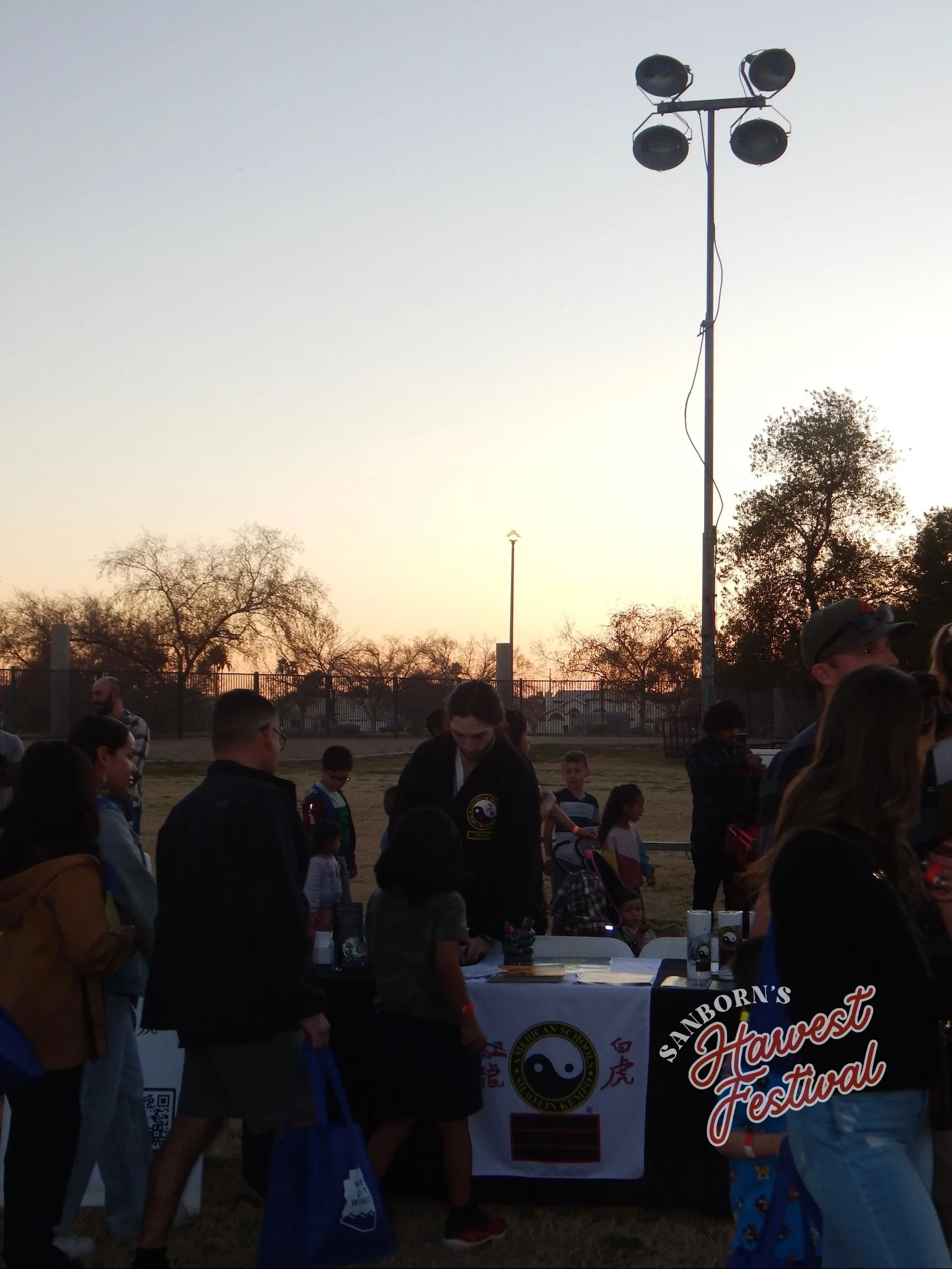 Group of people gathered around a table at an outdoor festival during sunset with trees in the background, a tall lighting pole, and a sign that reads "Sanborn's Harvest Festival".
