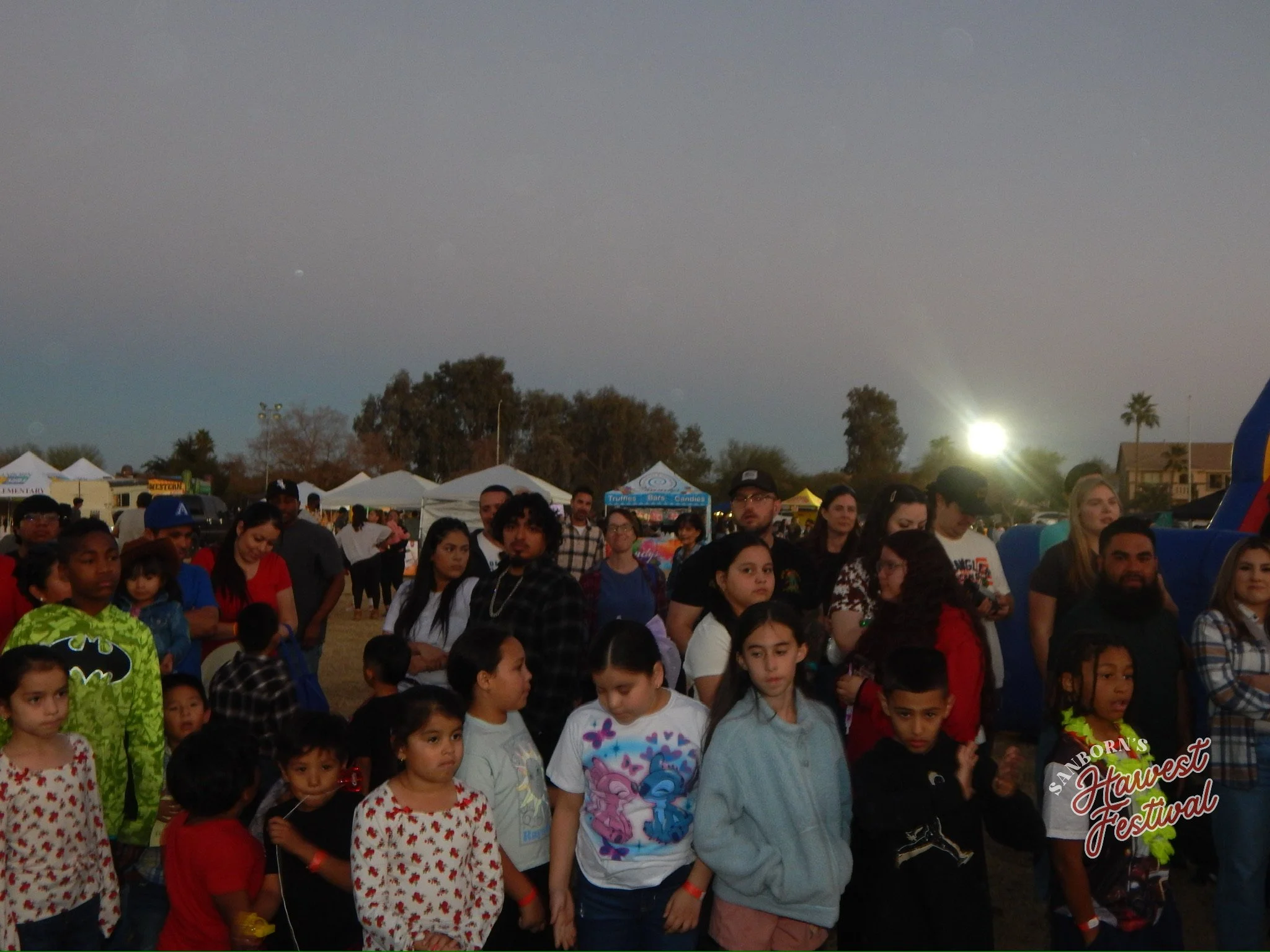 A large group of children and adults gathered outdoors at a festival during the evening, with tents, trees, and a bright moon in the sky.
