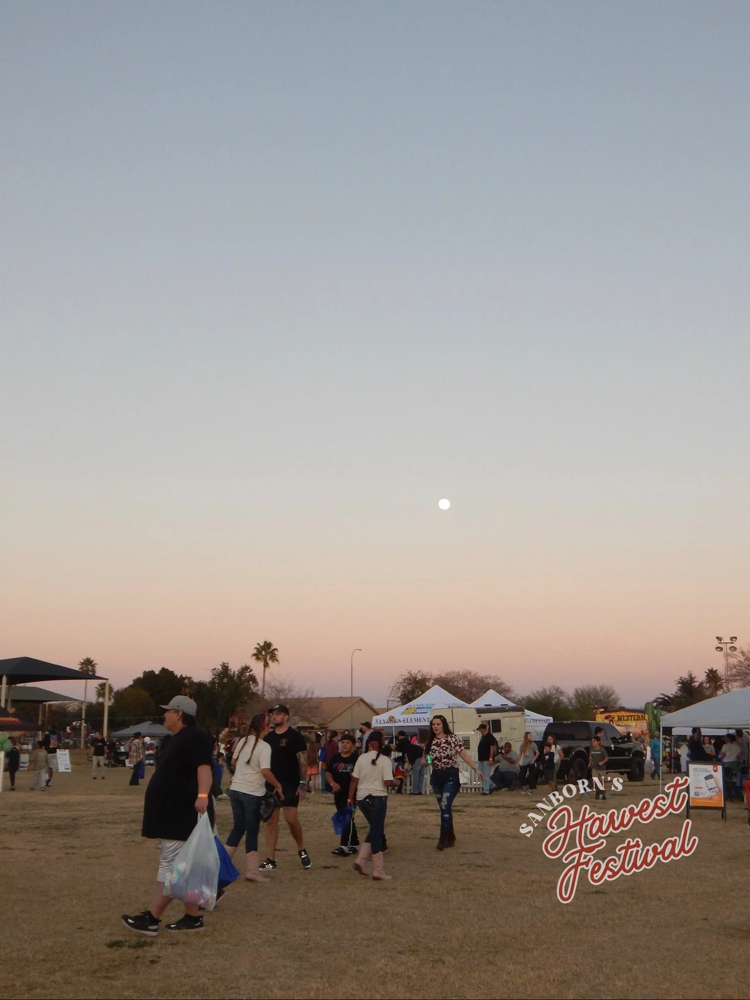 People walking at an outdoor fall festival with tents and signs, under a sky with the setting sun.
