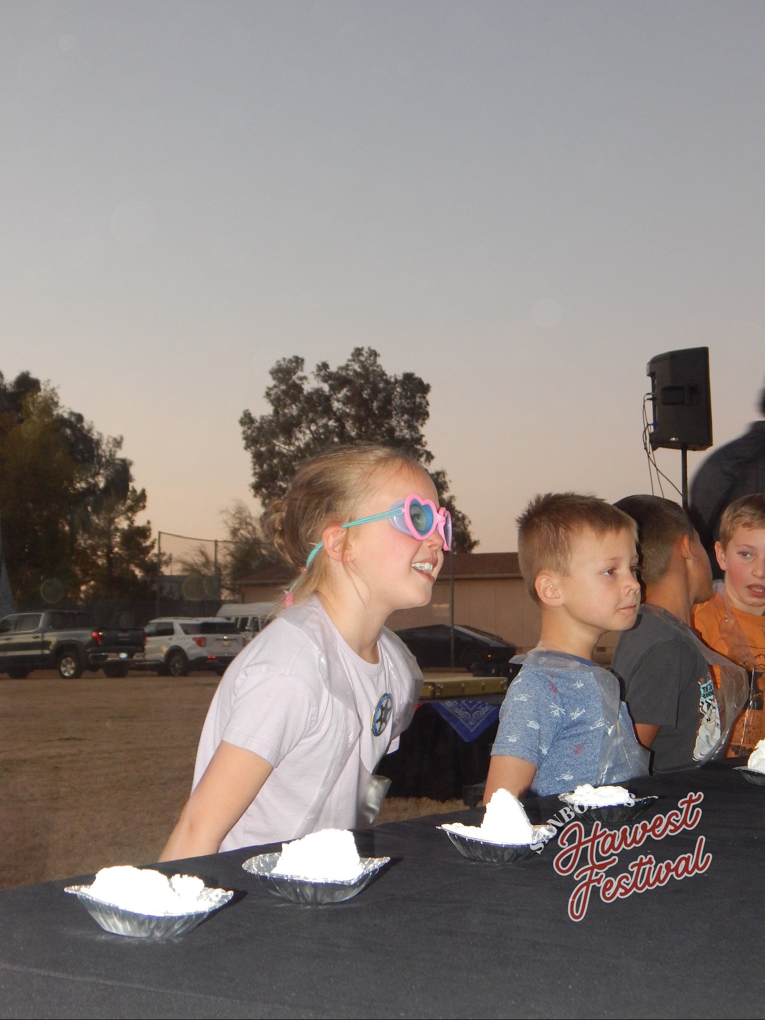 Children at an outdoor harvest festival enjoying desserts, with a table displaying three silver bowls of ice cream, trees, parked cars, and a speaker in the background.