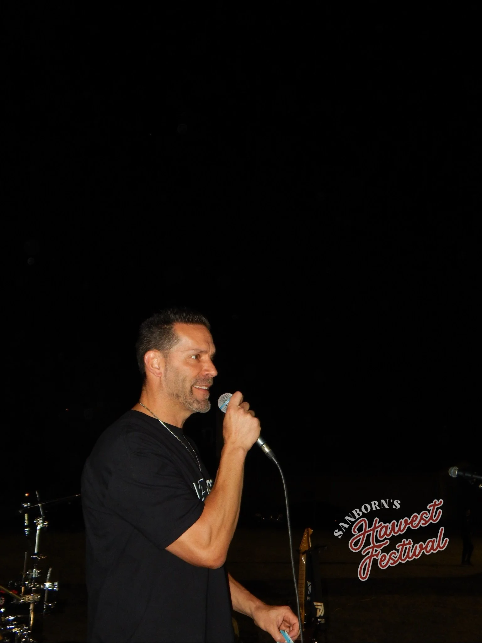 A man with short dark hair and a beard, holding a microphone and speaking at the Sanborn's Harvest Festival during nighttime.