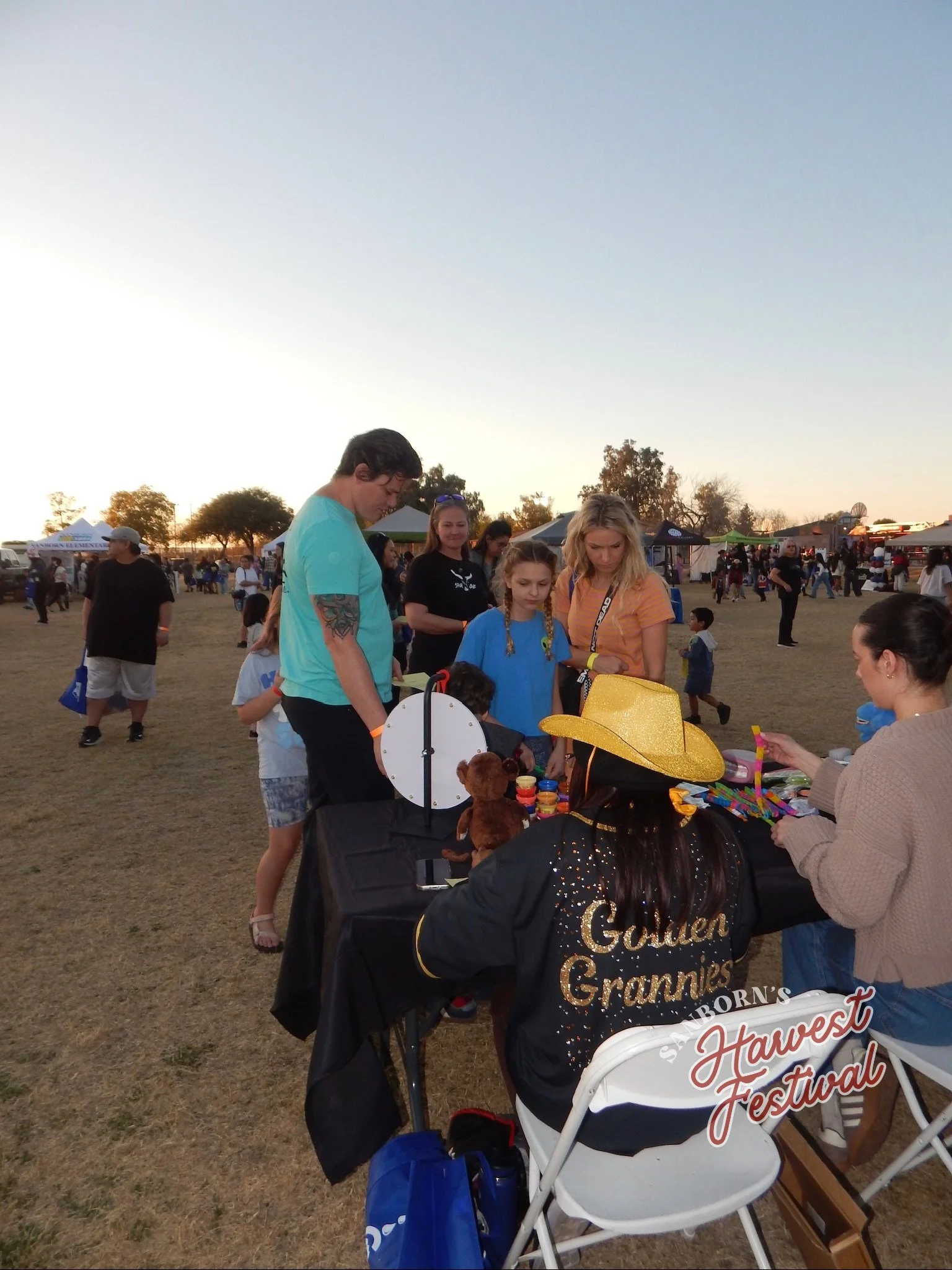 People at an outdoor festival, some shopping or browsing at a booth, with tents and other festival-goers in the background during sunset.