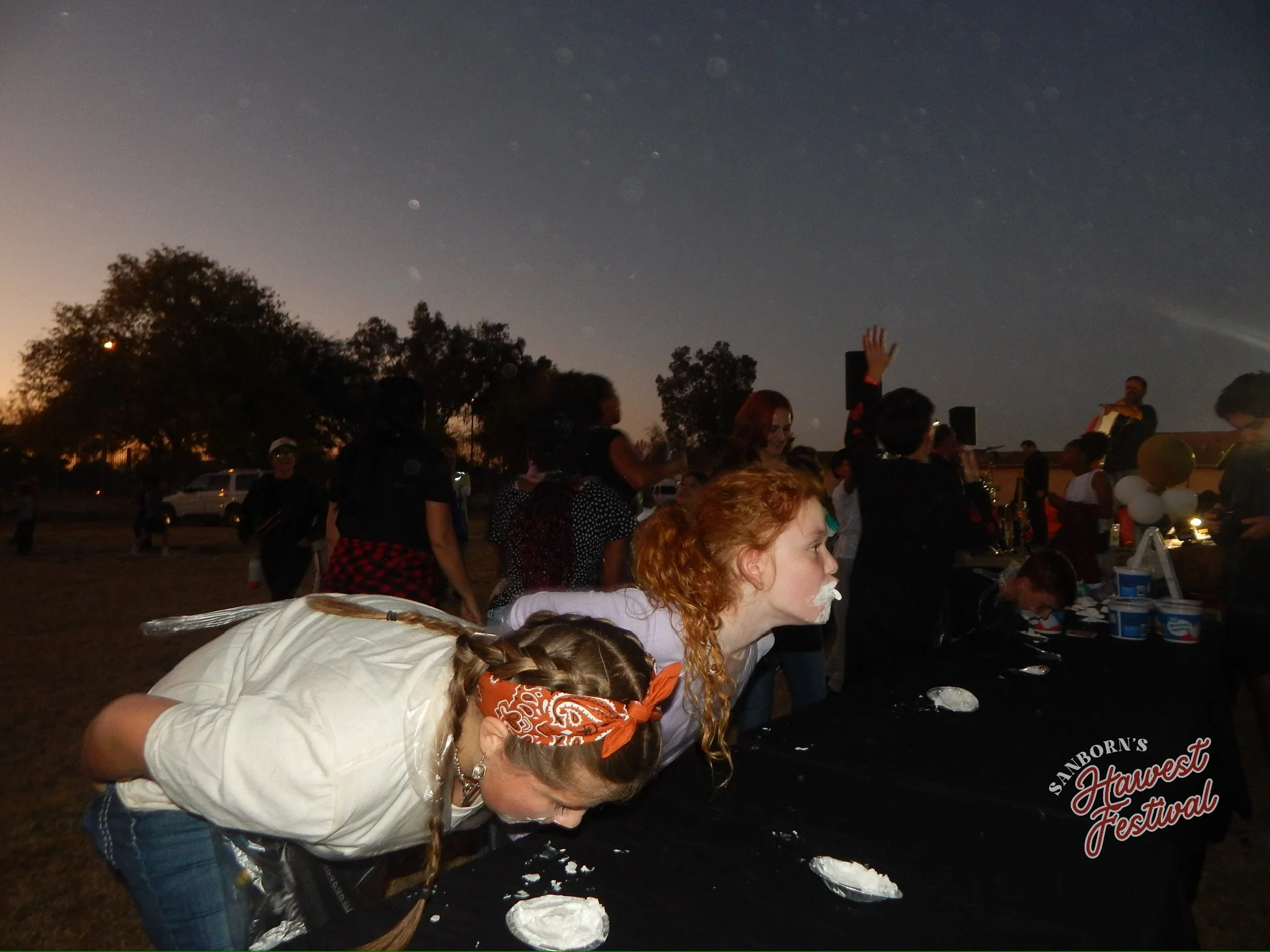People gathered outdoors at dusk, some with faces painted, participating in a festival or event with a food stand that has a sign reading 'Sanborn’s Harvest Festival.'