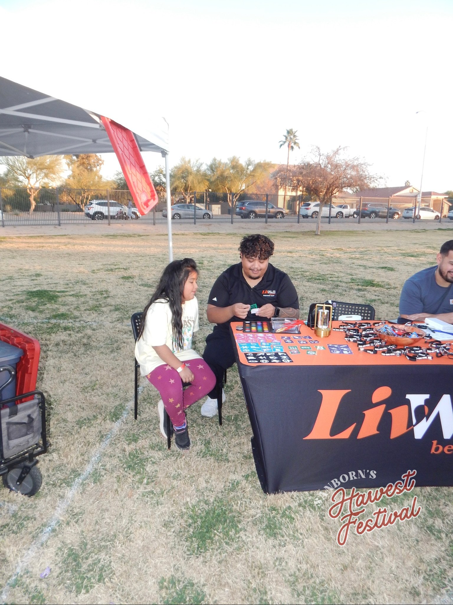 A young girl and a man sitting at a table outdoors at a harvest festival, with a black tablecloth, games, and prizes on the table, under a canopy tent, with cars and trees in the background.