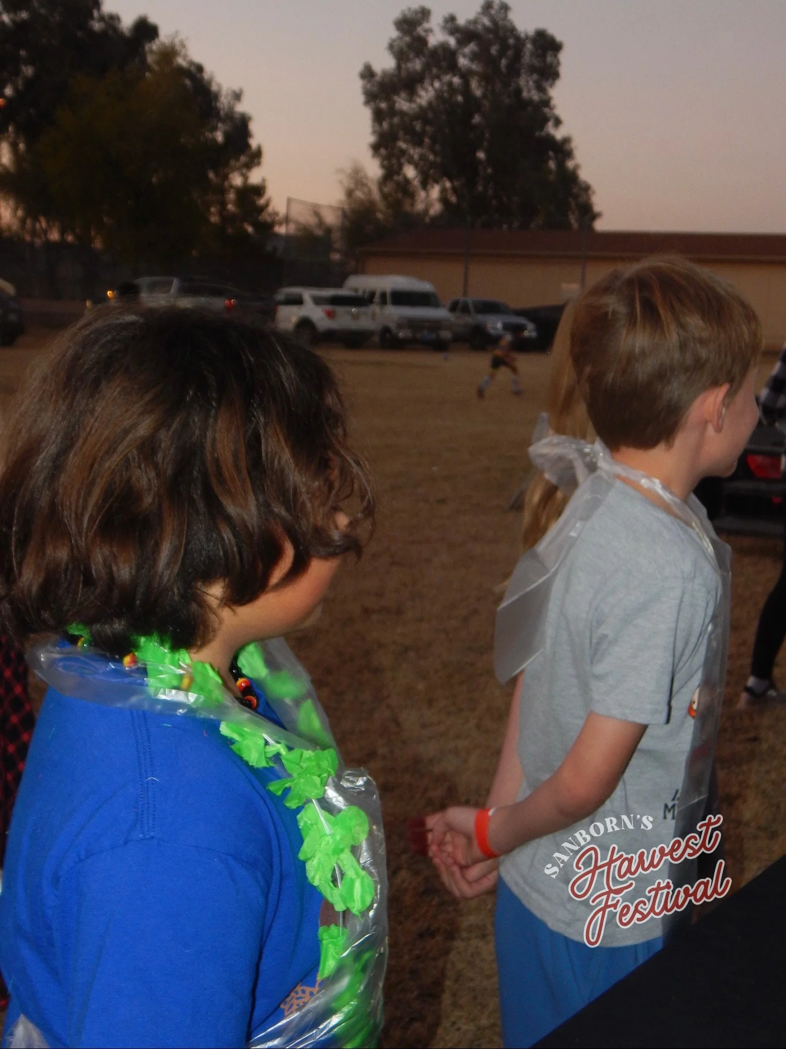 Close-up of children at an outdoor event during sunset, with a background of parked cars and trees.