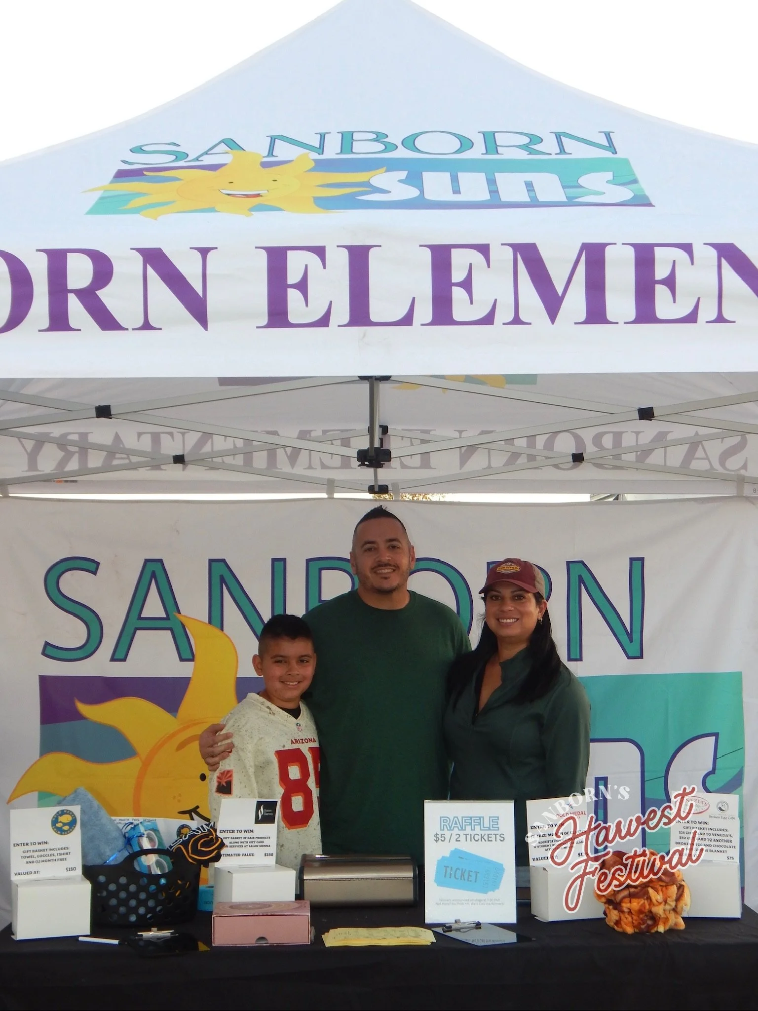 A man, woman, and boy standing behind a table at the Sanborn Harvest Festival. The table has raffle tickets, prizes, and donations, with a Sanborn Elementary School banner in the background.