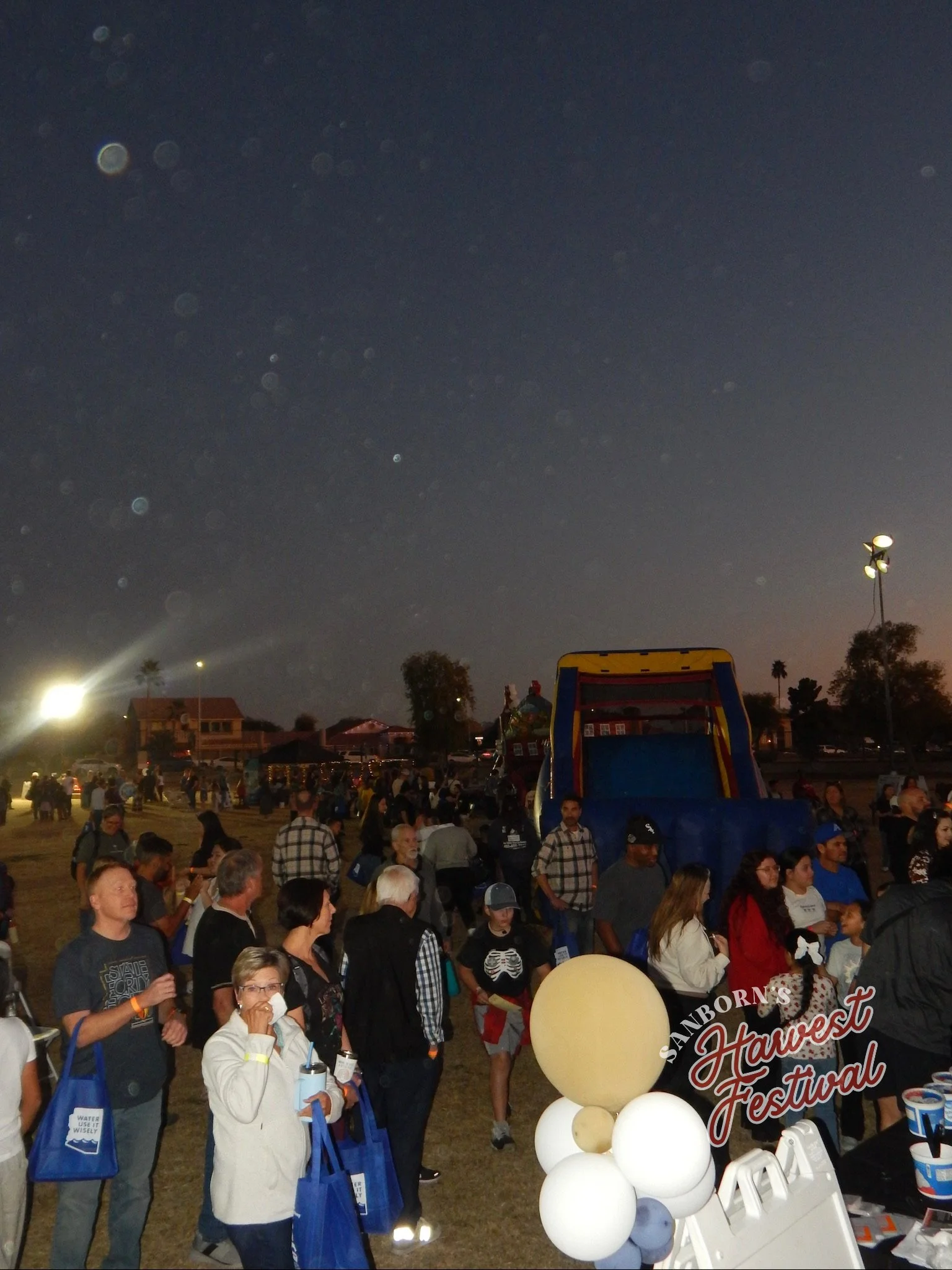 A large crowd gathered at an outdoor event during the evening. Many people are standing and walking around, with some holding balloons and blue tote bags. There is a sign that reads 'Sanborn's Harvest Festival.' In the background, there are trees, ho