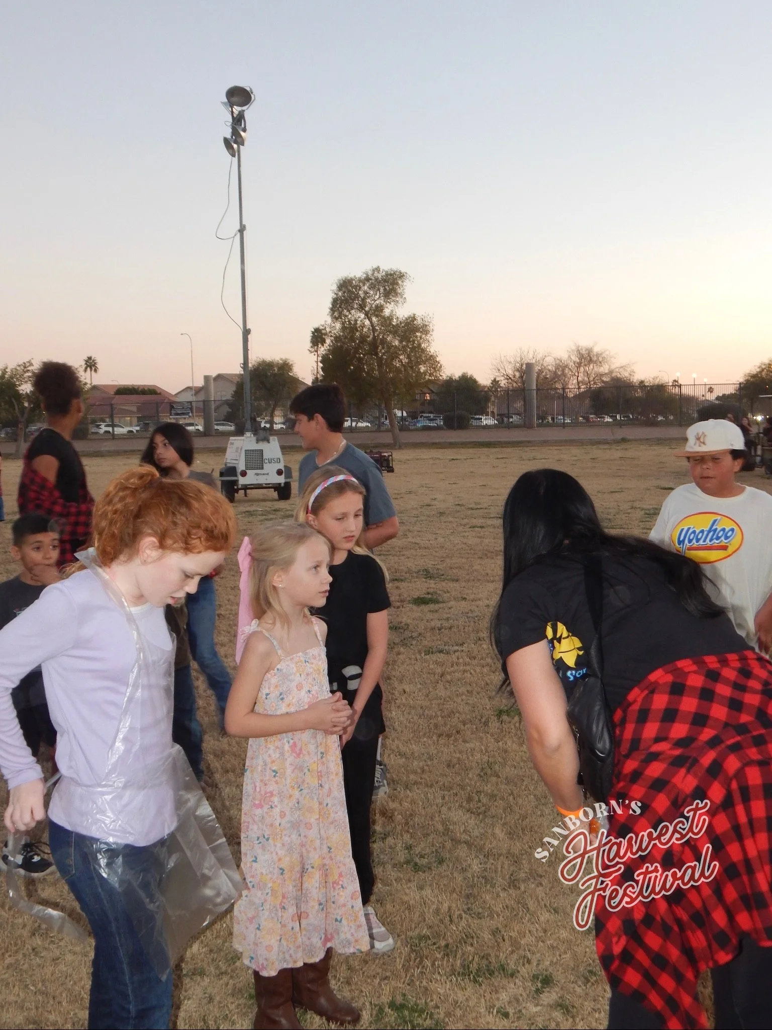 A group of children and adults gathered outdoors on a grassy field at sunset, participating in a community event called 'Sandborn's Harvest Festival,' with a tall light pole and trees in the background.