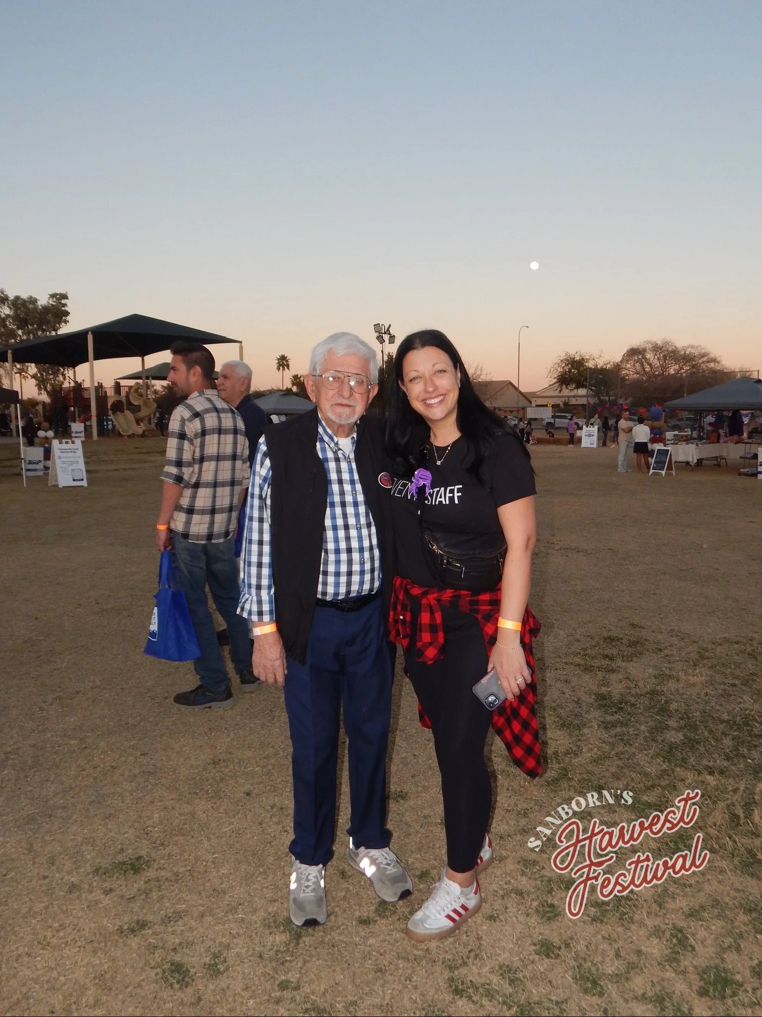 Two women standing together smiling at a harvest festival, with tents and people in the background during evening, one wearing a 'Volunteer Staff' shirt and the other in casual clothing.