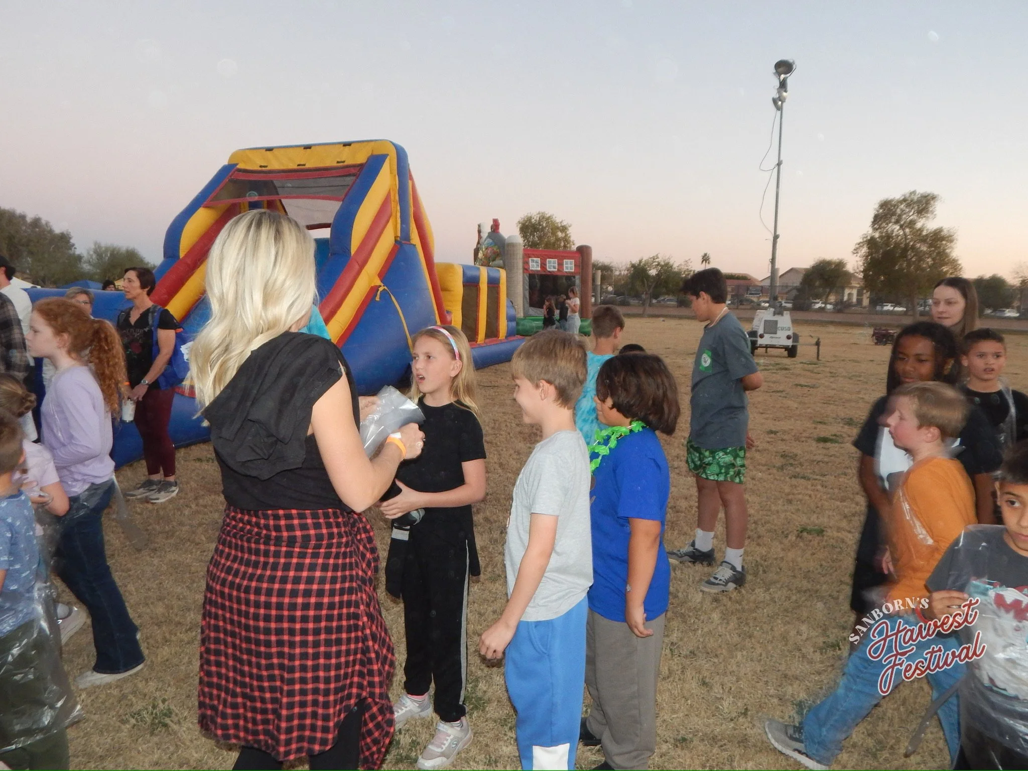 Children and adults gathered at an outdoor event with a colorful inflatable slide and a food stand in the background, during sunset in a park.