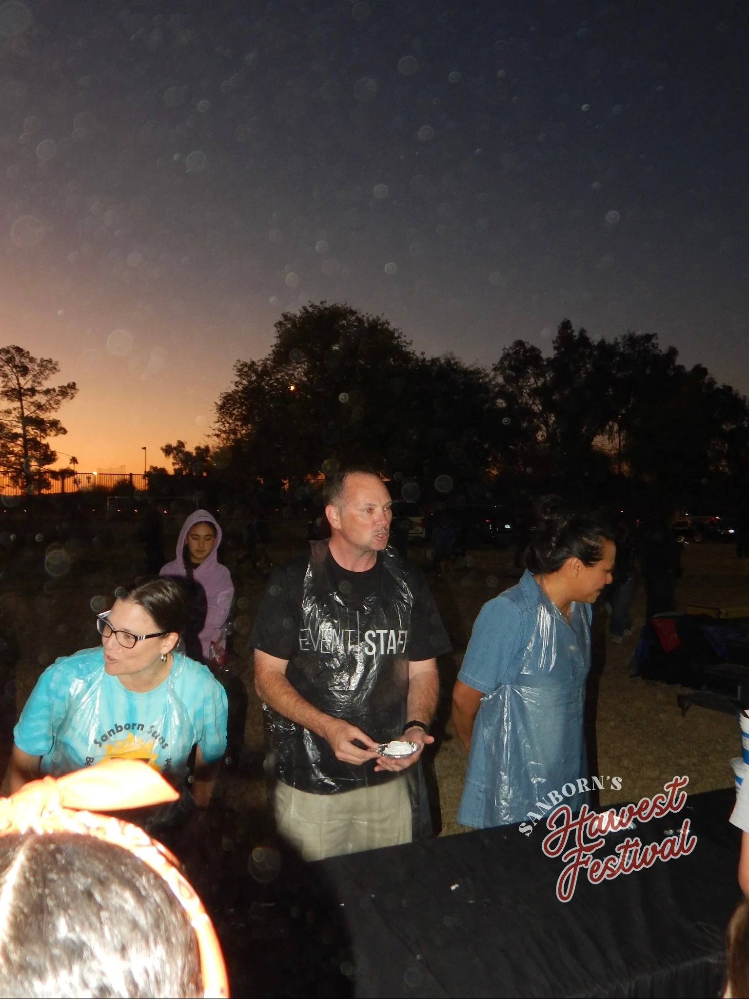 People gathered outdoors during a festival at dusk, with trees and the sky in the background. A man and several women are visible, some wearing rain ponchos. The festival appears to be called 'Sanborn's Harvest Festival'.