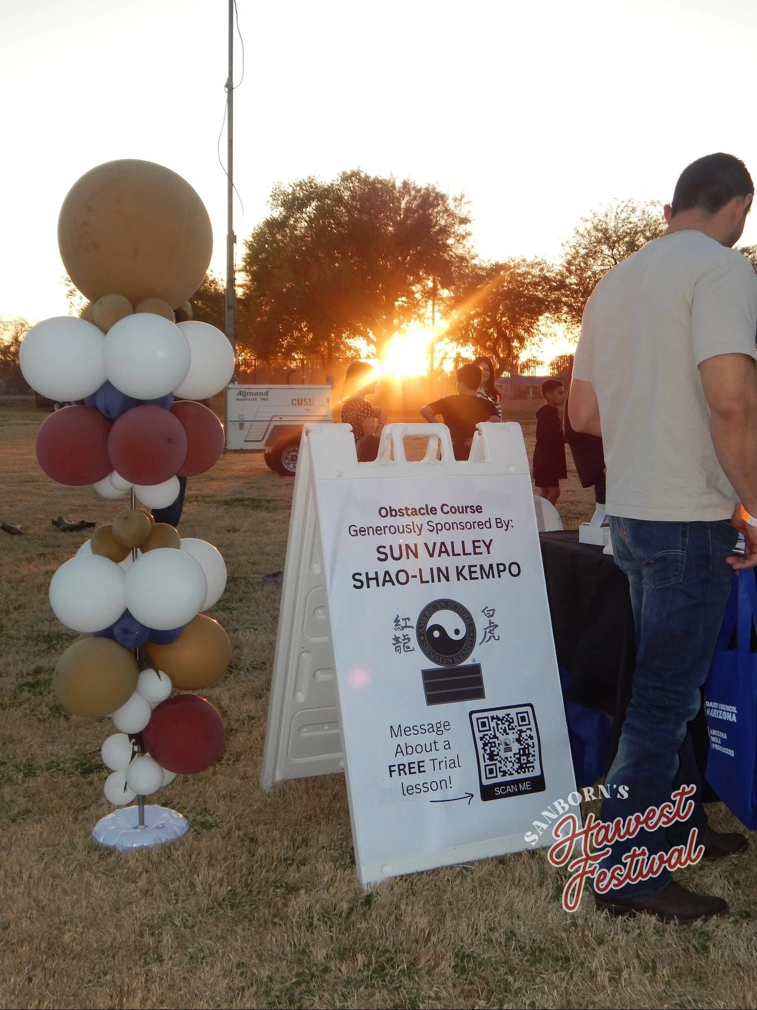 A balloon column with gold, white, blue, and maroon balloons standing on a grassy field near a sign at a festival during sunset. The sign invites people to take a free lesson about Shaolin Kempo karate, with a QR code for more information.