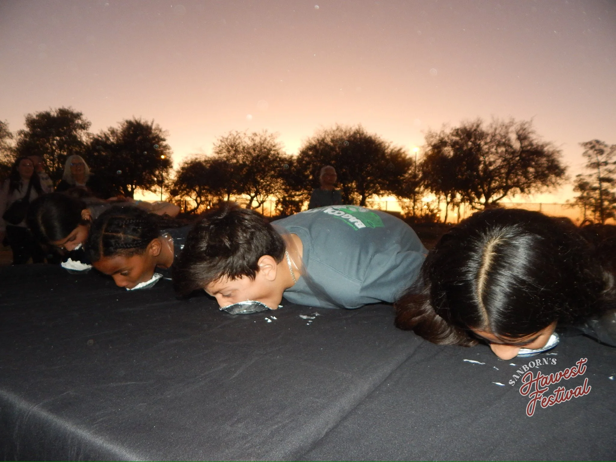 People with heads bowed, leaning over, on a black surface during sunset at an outdoor festival, trees in the background, some women standing.