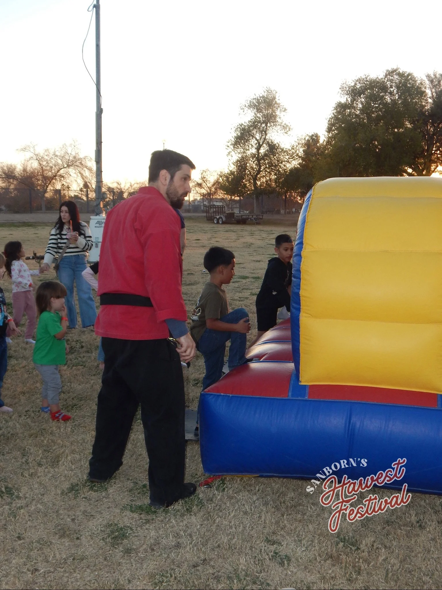 Group of children and adults at a fair or festival, standing near an inflatable bounce house. The setting is outdoors during sunset, with trees and a fence in the background.