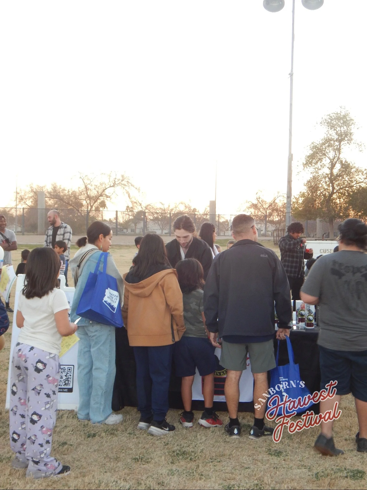 People gathered at an outdoor event, some standing in line at a booth with a sign reading "Sandborn's Harvest Festival." The scene is set in the evening with trees and a fence in the background.
