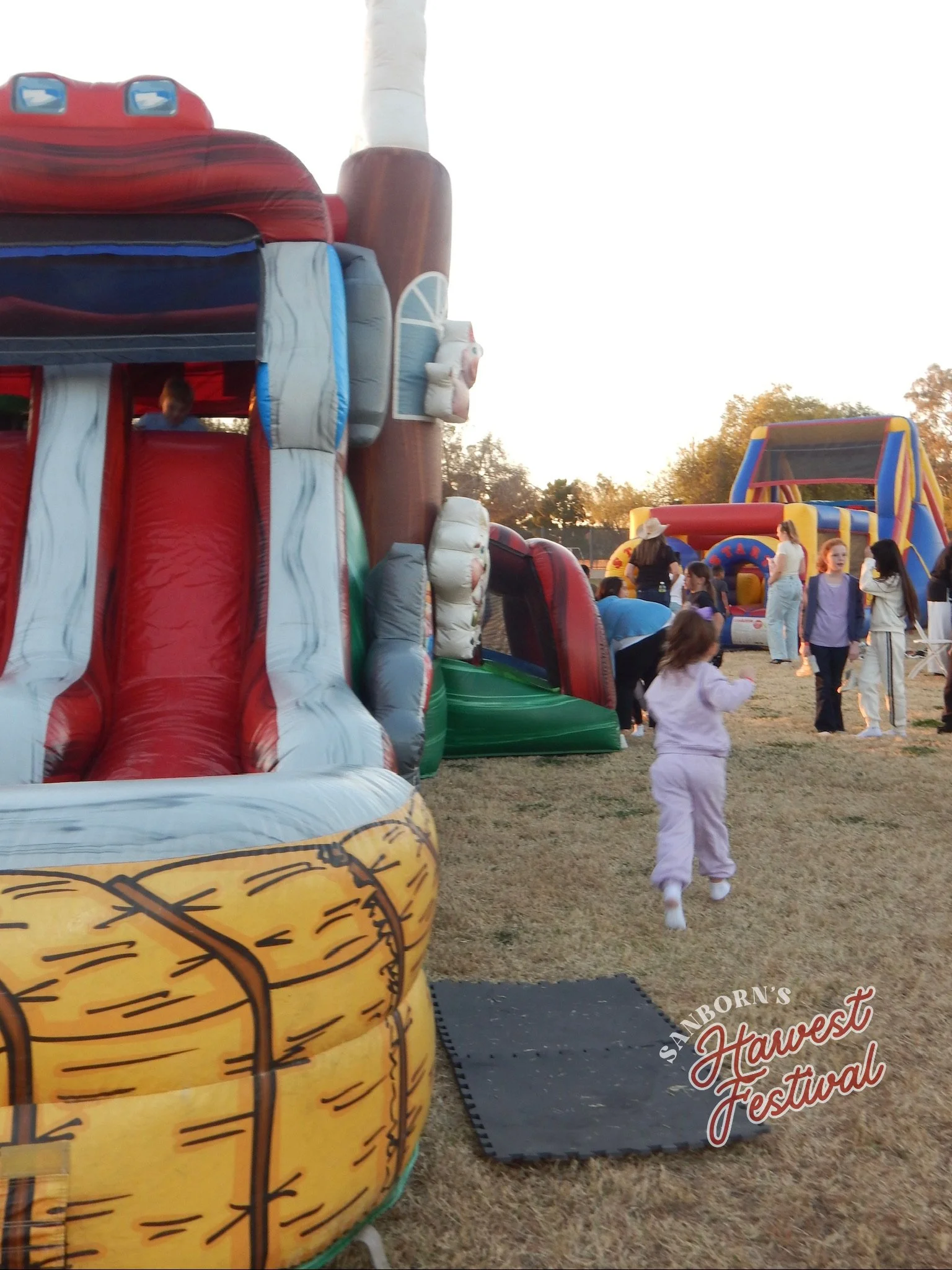 Inflatable bounce houses and slides at the Sanborn's Harvest Festival, with children playing and adults supervising outdoors during sunset.