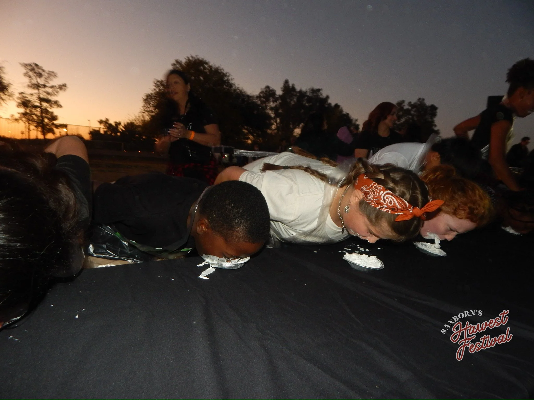 People participating in a pie-eating contest at an outdoor event during sunset, with trees and a fence in the background.