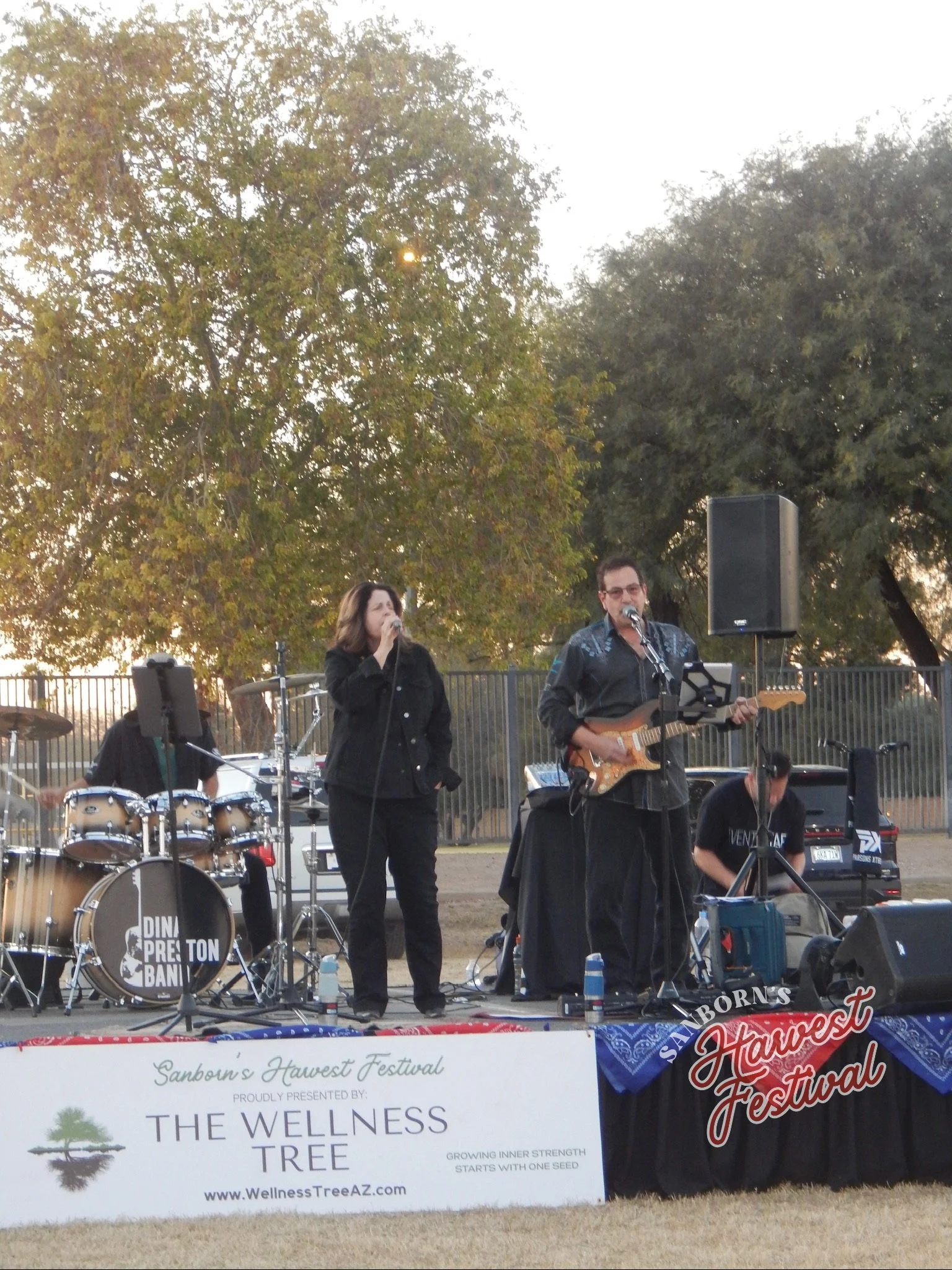A band performing on stage at Sanborn's Harvest Festival with a woman singing and a man playing guitar, surrounded by musical equipment, trees, and festival banners.