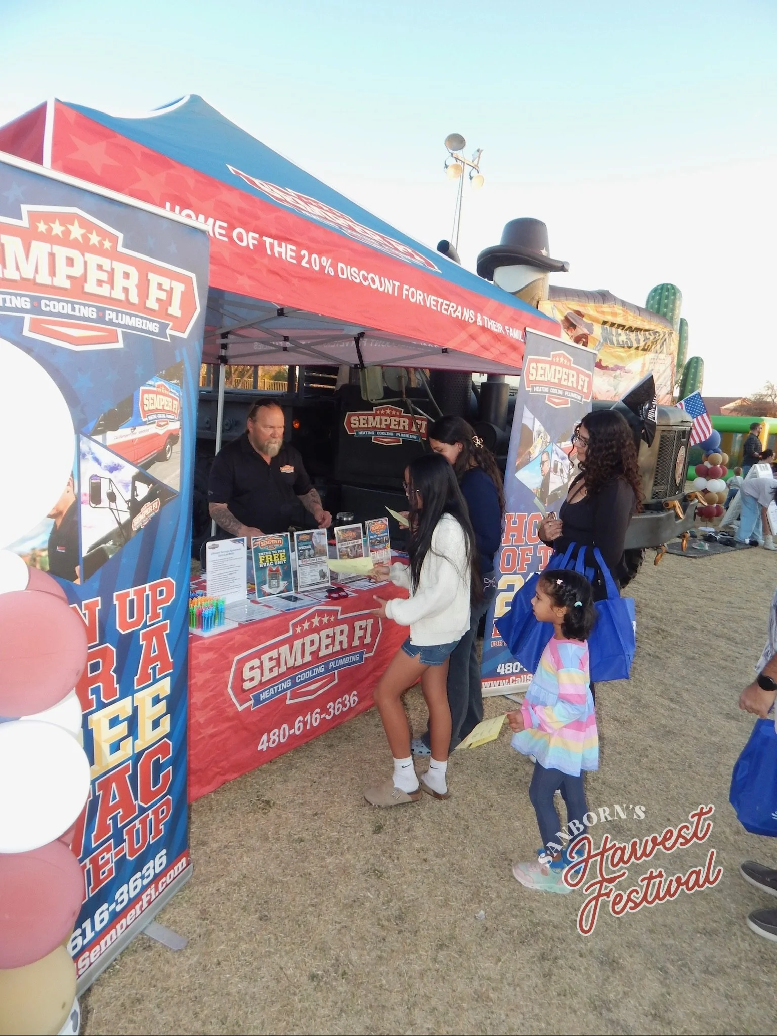 People gathered at a booth for Semper Fi Heating, Cooling & Plumbing at an outdoor festival, with children and balloons, and a decorated truck in the background.