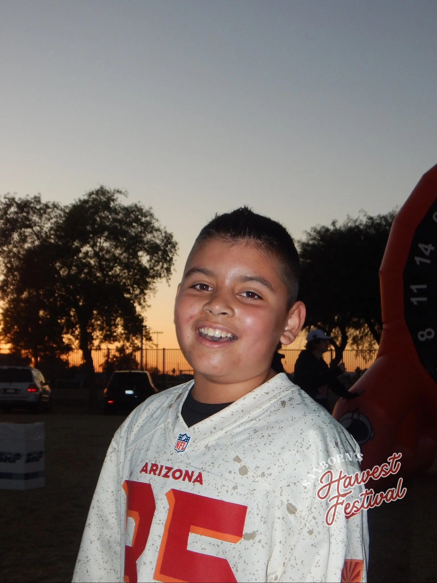 A smiling boy wearing a white Arizona Cardinals football jersey at an outdoor event during sunset, with trees and people in the background.