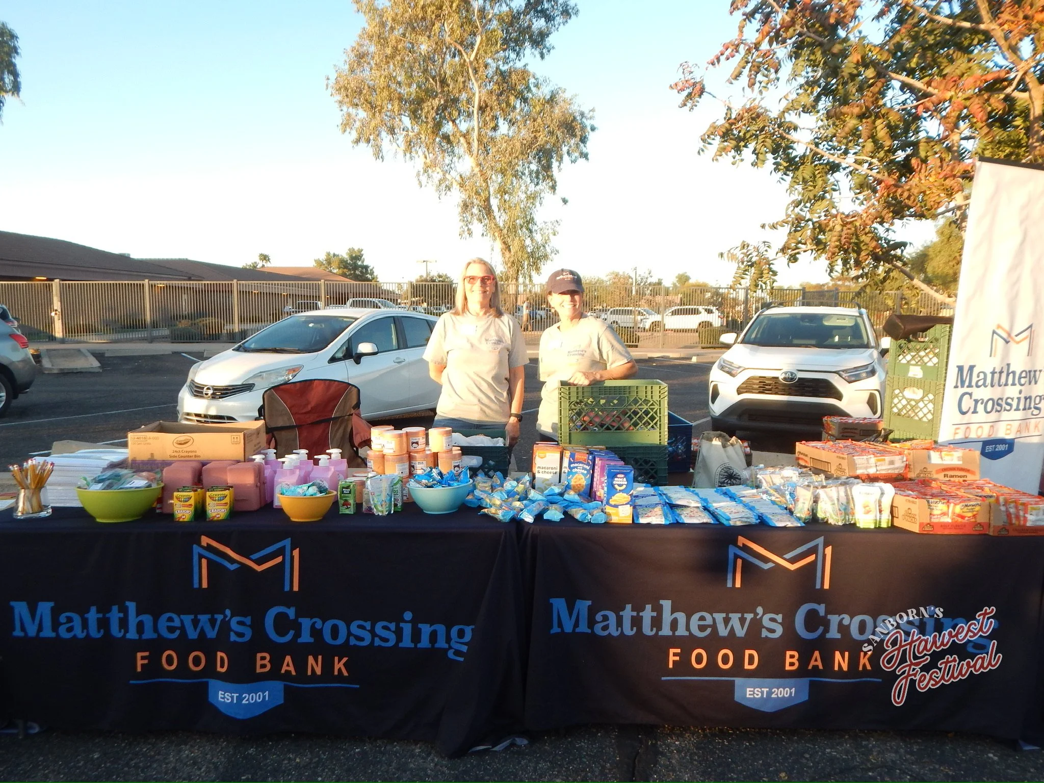 Two women standing behind a table with food items and supplies at a community food bank event, outdoors in a parking lot with trees and parked cars in the background, during daytime.