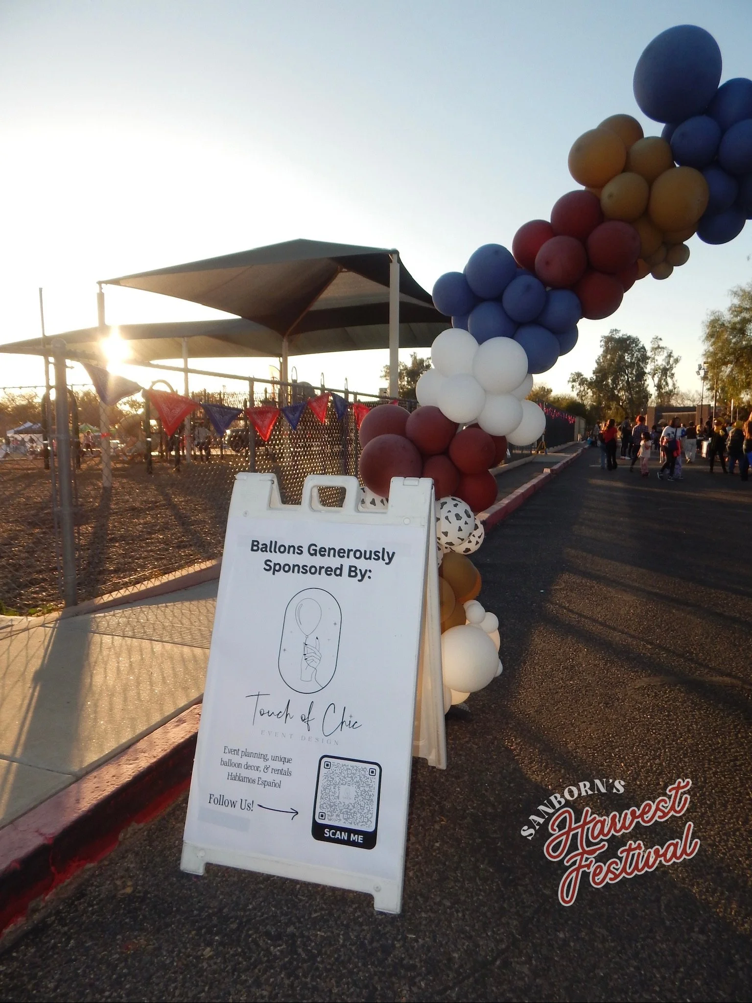 A display of balloons arranged in an arch at the Sanborn's Harvest Festival. There is a white sign with black text promoting the event, featuring a QR code for more information, and a QR code label at the bottom. The background shows people at the fe