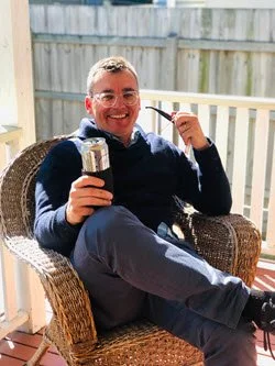 Man sitting outdoors on a wicker chair, smiling, holding a tumbler, with a pipe in his mouth, on a wooden deck with a fence in the background.