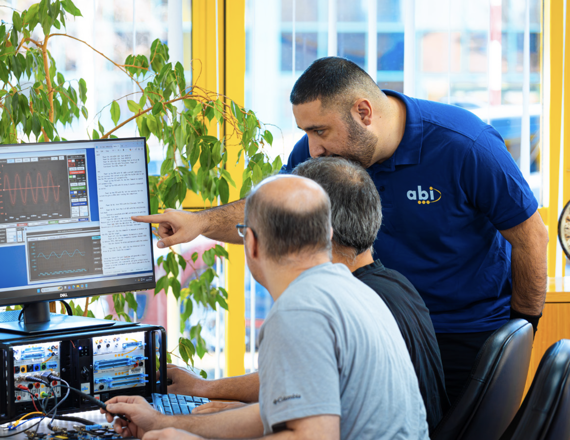 A man with a closely shaved hairstyle and beard wearing a blue polo shirt instructs several men seated at a desk, working on electronic devices and computers. They are in a brightly lit room with large windows, yellow walls, green plants, and an electronic testing or workbench with wires and circuit boards.