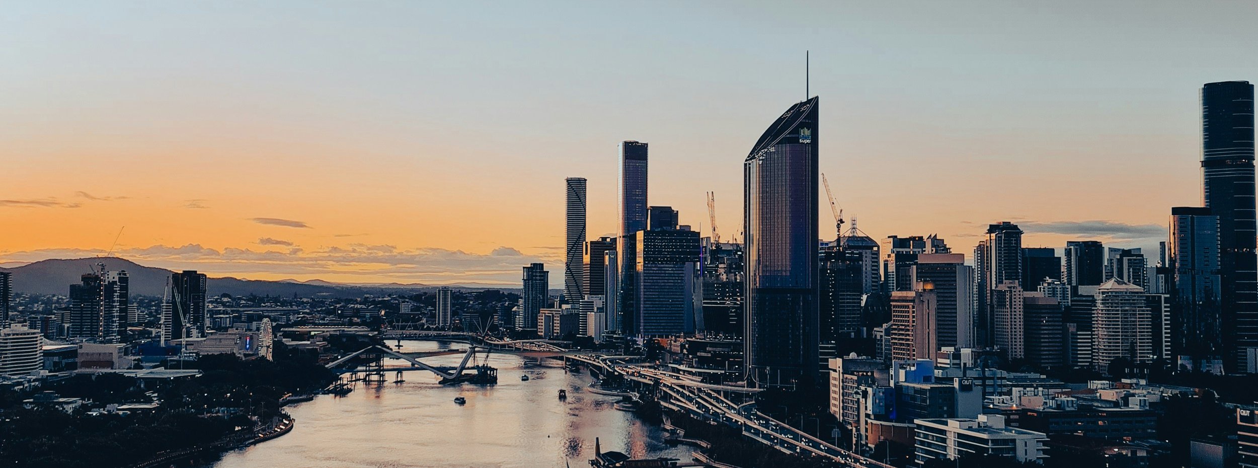 A city skyline at sunset with tall skyscrapers, a river running through the city, and mountains in the distance.