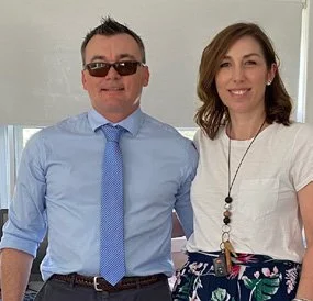 A man wearing sunglasses, a blue shirt, and a matching blue tie beside a woman in a white top and colorful skirt, both smiling.
