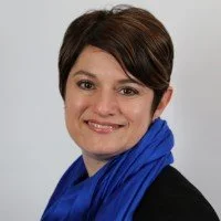 A woman with short brown hair smiling, wearing a black top and a blue scarf against a light gray background.