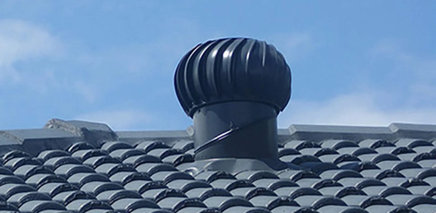 Close-up of a black, vented roof turbine on top of gray, curved rooftop tiles under a partly cloudy sky.
