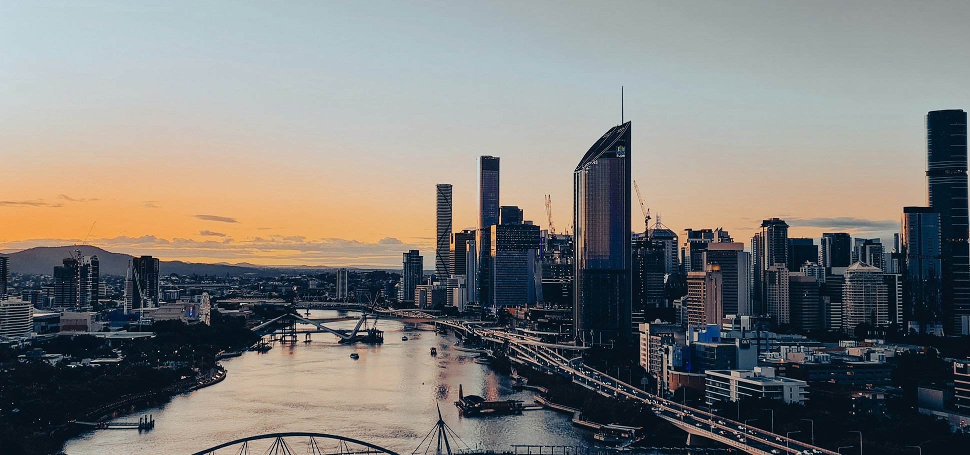 City skyline of Brisbane, Australia, with tall skyscrapers, a river in the foreground, and a sunset sky.