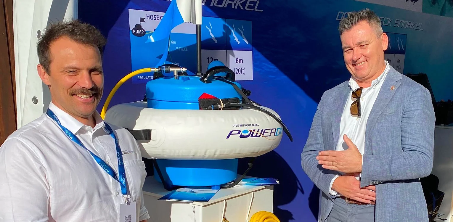 Two men smiling and standing beside a display of a compact blue and white underwater diving device or robot with the word "PowerD" on it, at an exhibition or conference.