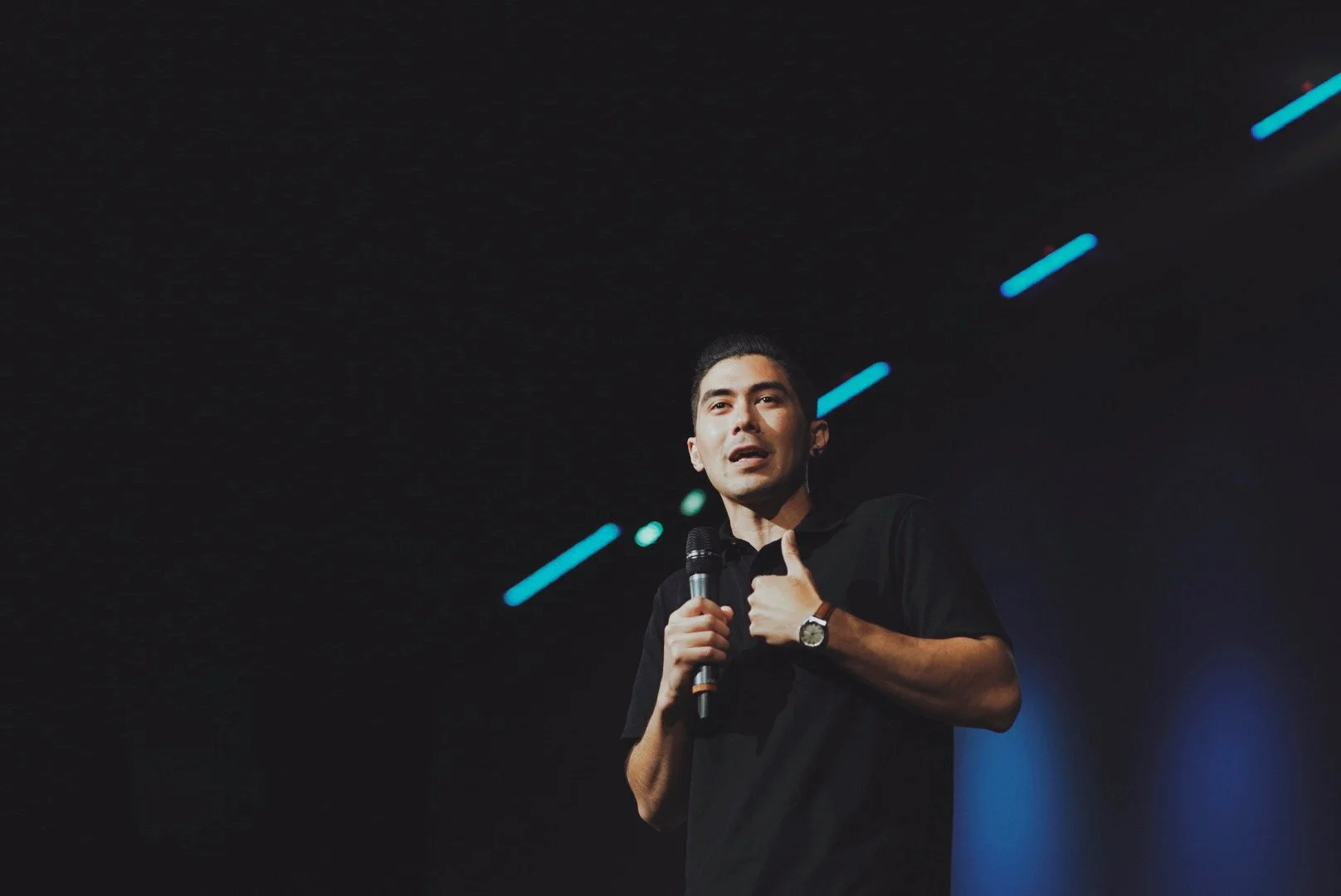 A man holding a microphone, standing on a stage with black background and blue lights.