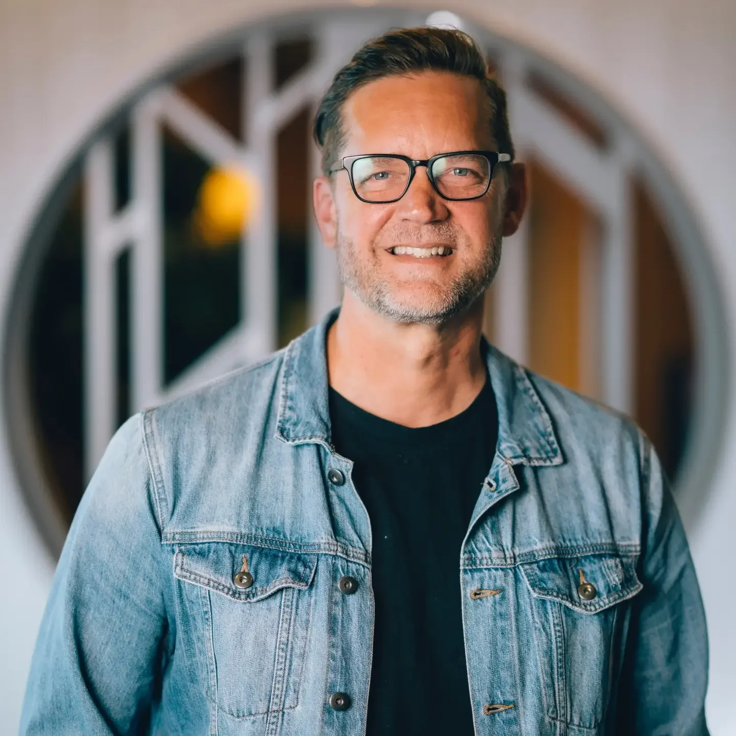 Smiling man with glasses and short dark hair, wearing a denim jacket and black shirt, standing in front of a round mirror with a white frame.