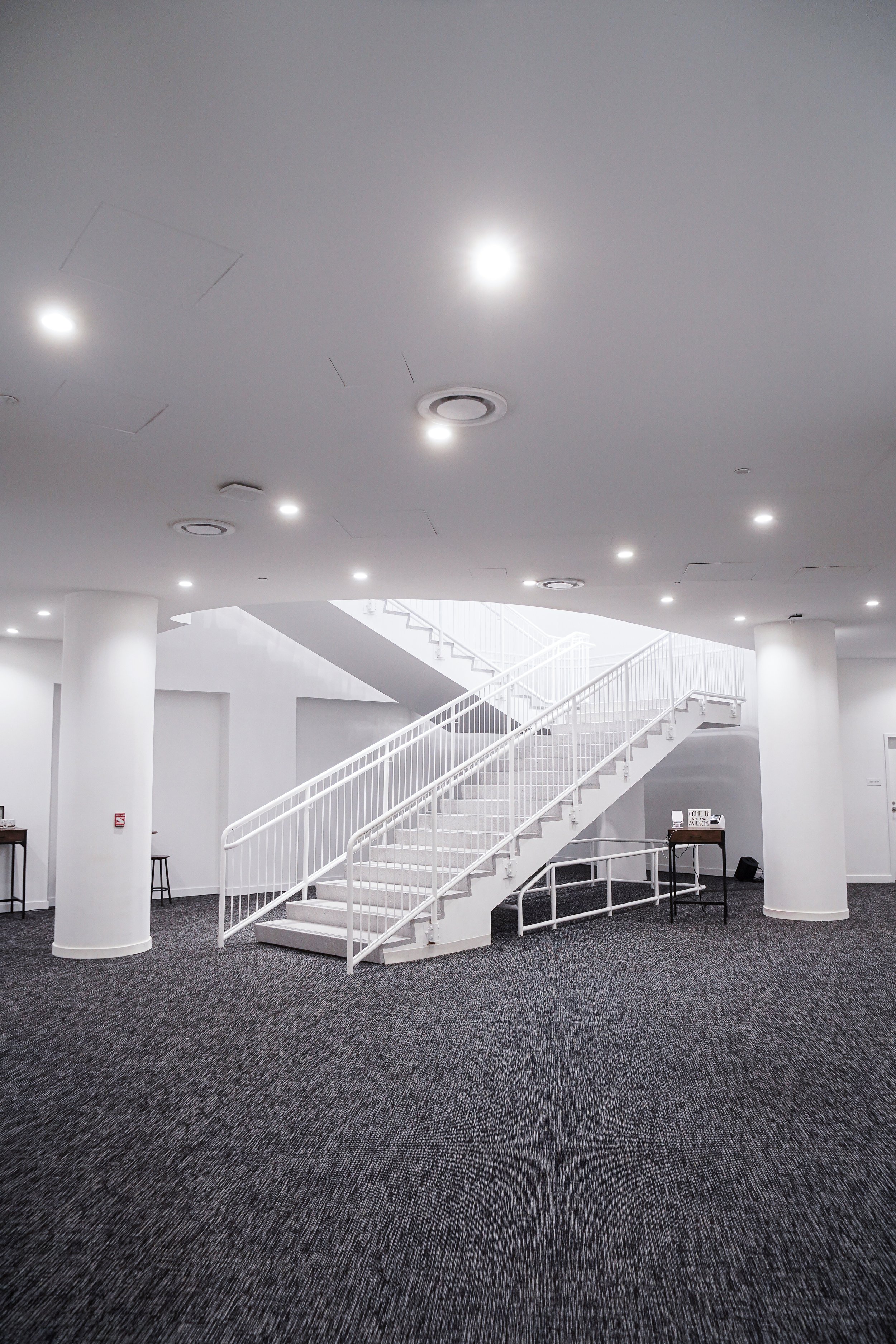 Interior of a modern building with white walls, a staircase with white railings, and a carpeted floor. Ceiling lights and air vents are visible.