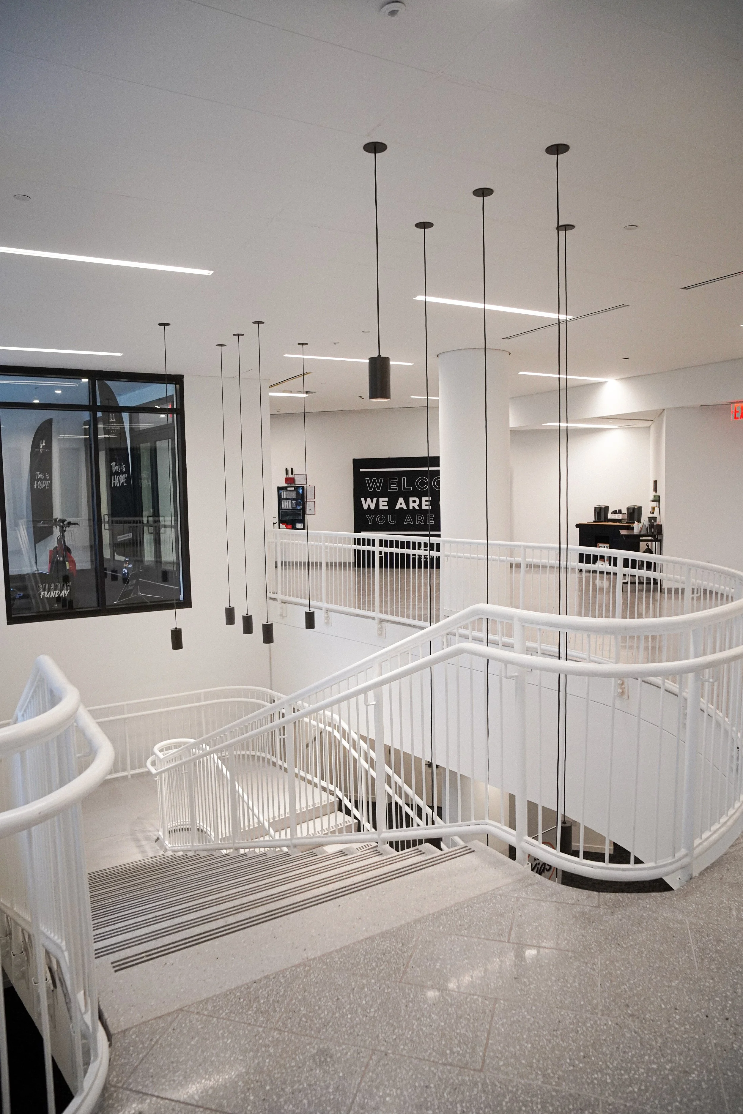 Interior view of a modern building with white walls and a staircase with white railings. Several hanging black pendant lights are suspended from the ceiling. In the background, there is a black sign with white text that reads 'WELCOME WE ARE YOU ARE'. There is also a black vending machine and a table with some items on it. The floor is covered with gray and white speckled tiles.