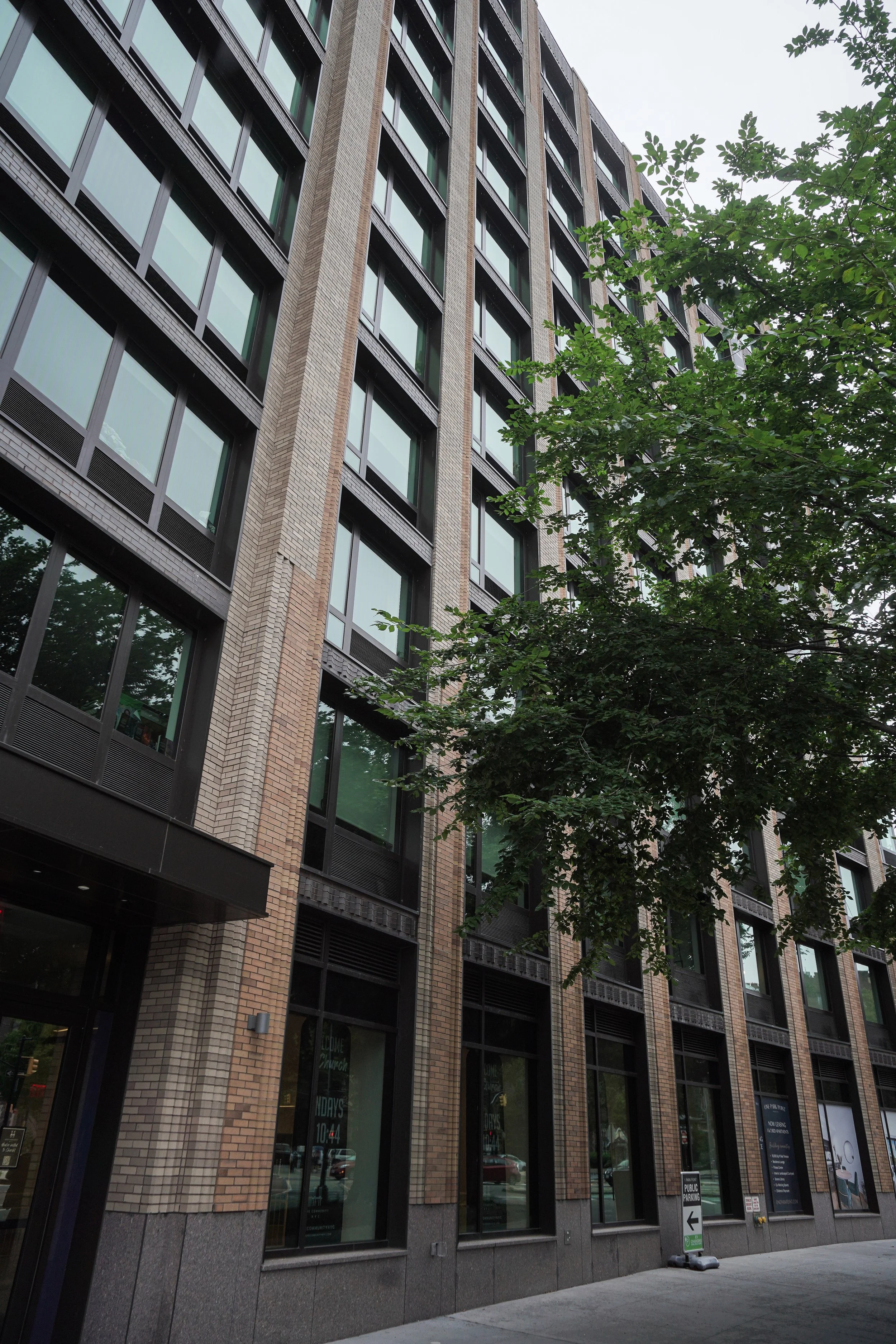 A multi-story modern building with large glass windows and brick facade, partially obscured by a tree with green leaves.