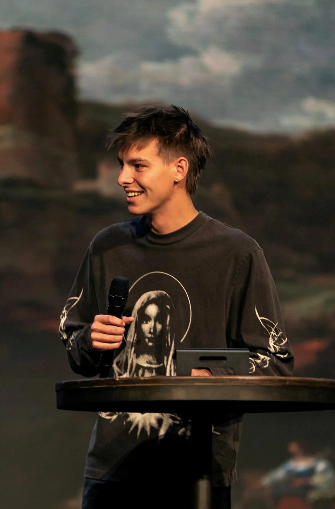 Young man with short, spiky hair smiling, holding a microphone, wearing a black sweatshirt with graphic print of a woman on the front, standing behind a lectern with a tablet, against a blurry background, possibly at an indoor event or presentation.