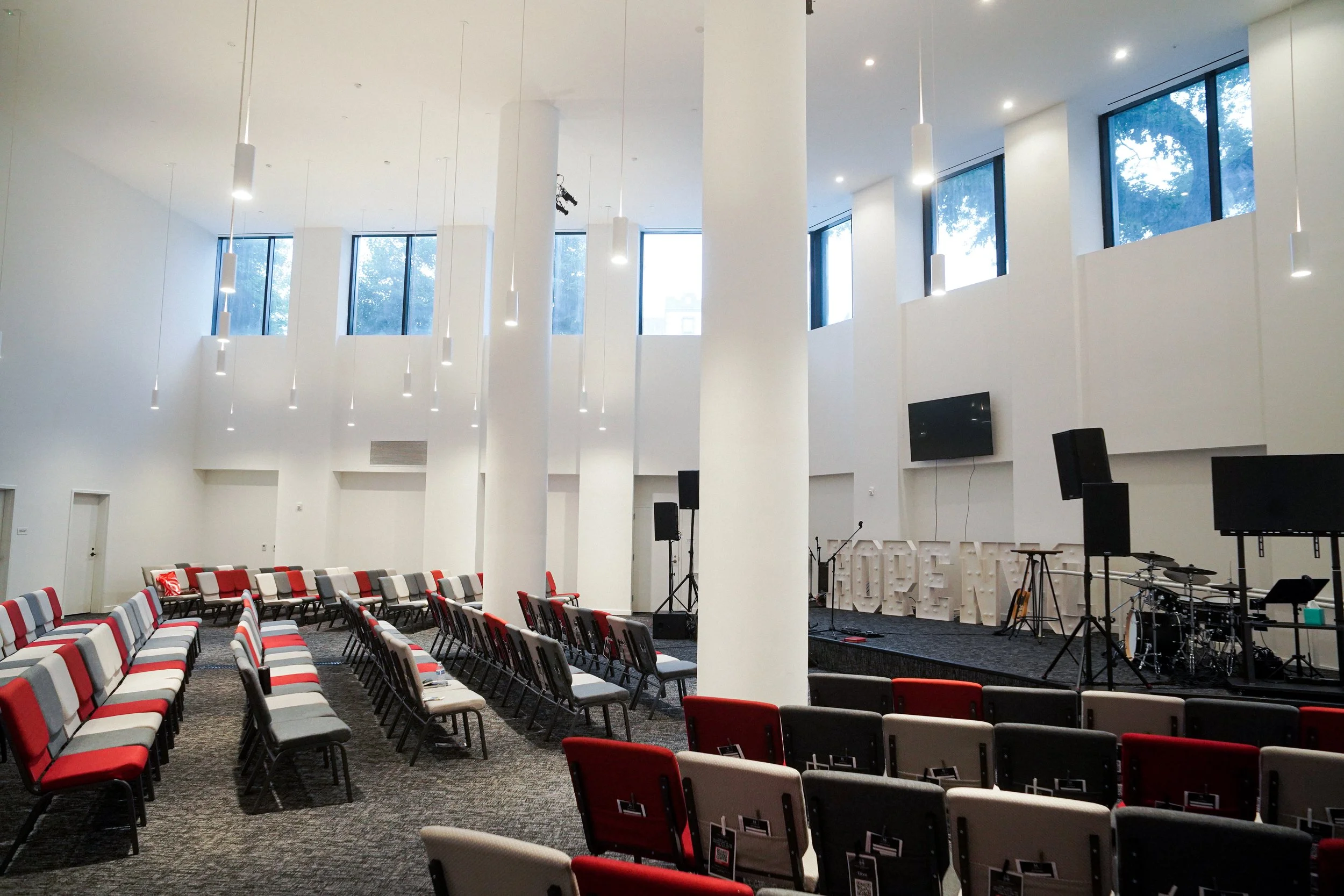Empty event space with rows of red, gray, and beige chairs facing a stage with musical instruments and large screens. Modern white walls and large windows with trees outside.